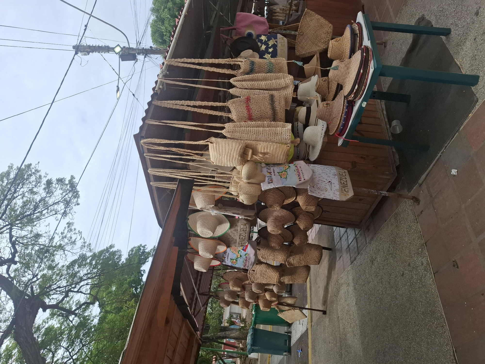 The image captures an outdoor market stall, likely in El Valle del Espíritu Santo, Venezuela, under a bright, clear daytime sky. To the left, large green trees and a utility pole with multiple power lines are visible against the light sky. The market stall, constructed of wood, features a prominent display of local handicrafts.
Numerous straw and woven items are offered for sale. Rows of handcrafted bags and purses, made from natural fibers, hang from the stall's frame. Some bags exhibit distinctive horizontal stripes in red, blue, or green. Various straw hats, many with colored trims, are neatly arranged on stands and shelves. Smaller woven baskets and decorative pieces also contribute to the display.
Among the items, a light-colored fabric bag or banner stands out, clearly displaying the word "VENEZUELA" alongside a colorful map. The text "LA MARGARITA" is also partially visible on this item, providing a geographical clue consistent with the given location context. Other merchandise includes a pink fabric bag and a dark blue bag adorned with light-colored patterns, possibly depicting sea turtles, on a higher shelf.
No individuals are present in the frame. The paved ground or sidewalk in front of the stall indicates an urban or semi-urban setting, with a green wheeled trash bin visible further down the street. The scene depicts a typical craft market, suggesting an activity of commerce and tourism, with the clear skies indicating fair weather.