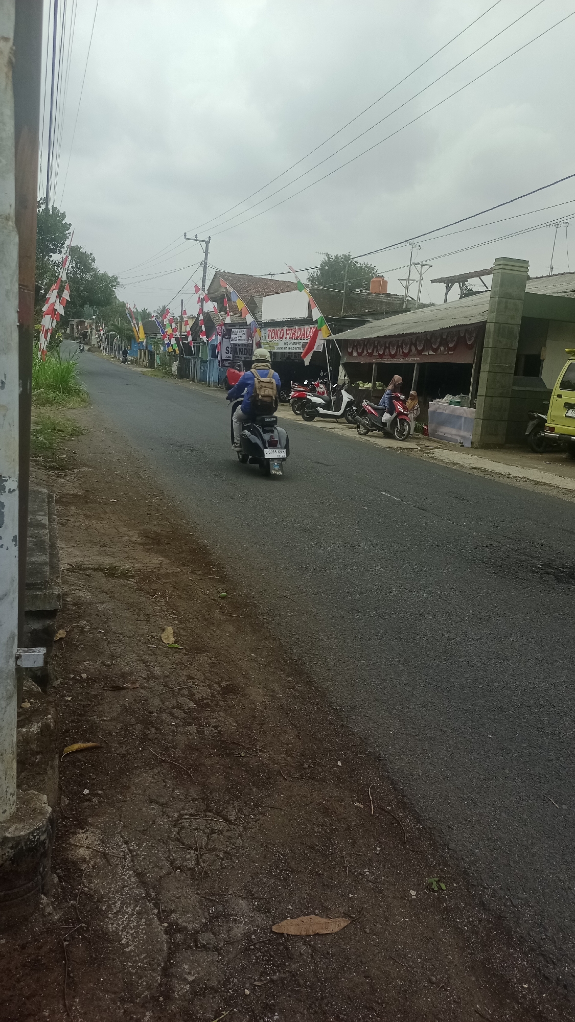 The image depicts a street scene in Karyamandala, Indonesia, under an overcast sky. The road surface is asphalt, showing some signs of wear and tear. On the left side of the road, there is a concrete structure, possibly a wall or curb, with a weathered appearance. Beyond this, greenery and trees are visible.
The right side of the road features several buildings, including a shop with a sign that reads "TOKO FIADAVA." In front of the shop, there are several motorcycles parked, and two people are visible, one sitting on a red motorcycle and another standing. A yellow vehicle, possibly a truck or van, is parked further down the road.
The street is decorated with numerous Indonesian flags and banners, suggesting a celebratory or patriotic event. A person is riding a dark-colored scooter in the foreground, facing away from the camera, wearing a blue jacket and a backpack. Another scooter is parked in the middle ground. The overall impression is a quiet, potentially residential or commercial street preparing for or participating in a local event.