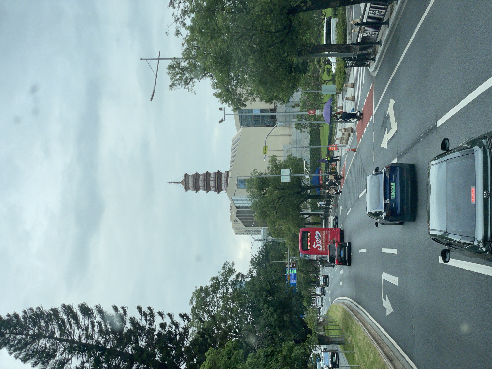 The image shows a street scene in Haizhu District, China, with traffic moving on a multi-lane road. A tall pagoda-style building is visible in the background, alongside modern buildings. Trees line the street, partially obscuring the view of the buildings. The sky is overcast. Traffic includes cars and a red bus. A crosswalk with pedestrians is present on the right side of the road.