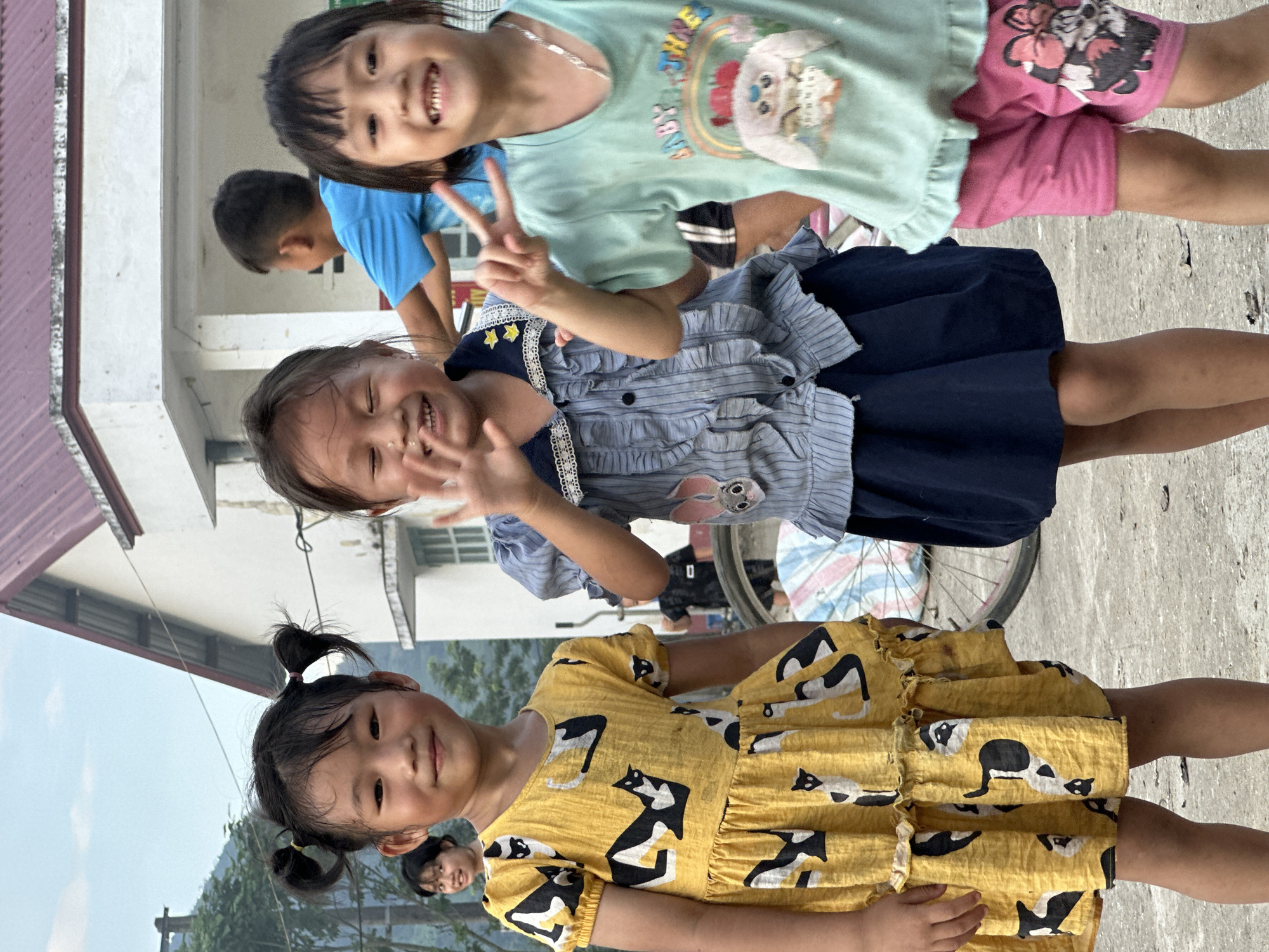The image displays an outdoor scene in Gia Hội, Vietnam, featuring three young girls in the foreground. From left to right: The first girl has dark hair styled in two pigtails with yellow hair ties and is wearing a yellow short-sleeved dress with black and white abstract cat patterns. She is smiling and looking directly at the camera. The second girl has dark hair and is wearing a blue and white striped collared shirt with ruffles and a dark blue skirt. A gray rabbit patch with pink ears is visible on her left chest area. Her eyes are closed in a wide smile, and her right hand is raised in a partial waving gesture. The third girl has dark, shoulder-length hair and is wearing a light green short-sleeved t-shirt with a graphic depicting a white cartoon character, a rainbow, and the text "BABY THREE." She is also wearing pink shorts with a Minnie Mouse graphic on the left leg and a silver necklace. She smiles broadly at the camera with her left hand raised, forming a peace sign.
In the background, on the far left, a woman with dark hair is partially visible, smiling. Behind the central girls, a boy wearing a blue t-shirt is partially visible, facing away from the camera. The ground is a light-colored paved surface. A white building with a dark reddish-brown corrugated metal roof is prominent in the background. Green trees and distant mountains are visible under a pale blue sky on the left side of the frame. A bicycle wheel and part of its frame, partially covered by light-colored fabric, are visible behind the girls near the building. The scene is illuminated by natural daylight.