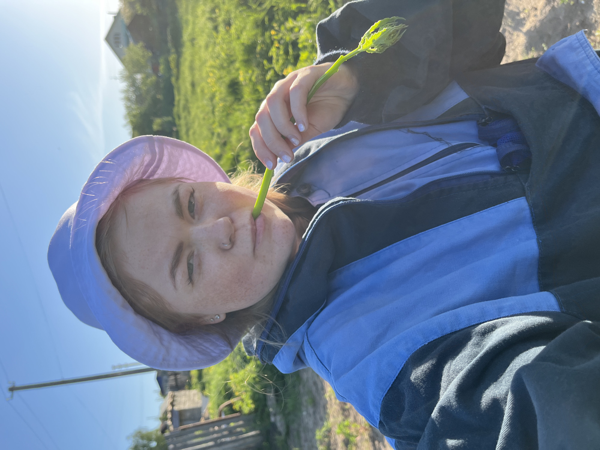 A young woman wearing a light purple sun hat and a blue and black jacket is holding a long green plant stem to her mouth. The stem has a cluster of leaves at the top. She has fair skin and freckles. The background shows a green field or grassy area with a house visible in the distance. The location is identified as the city of Ольховка, Russia.