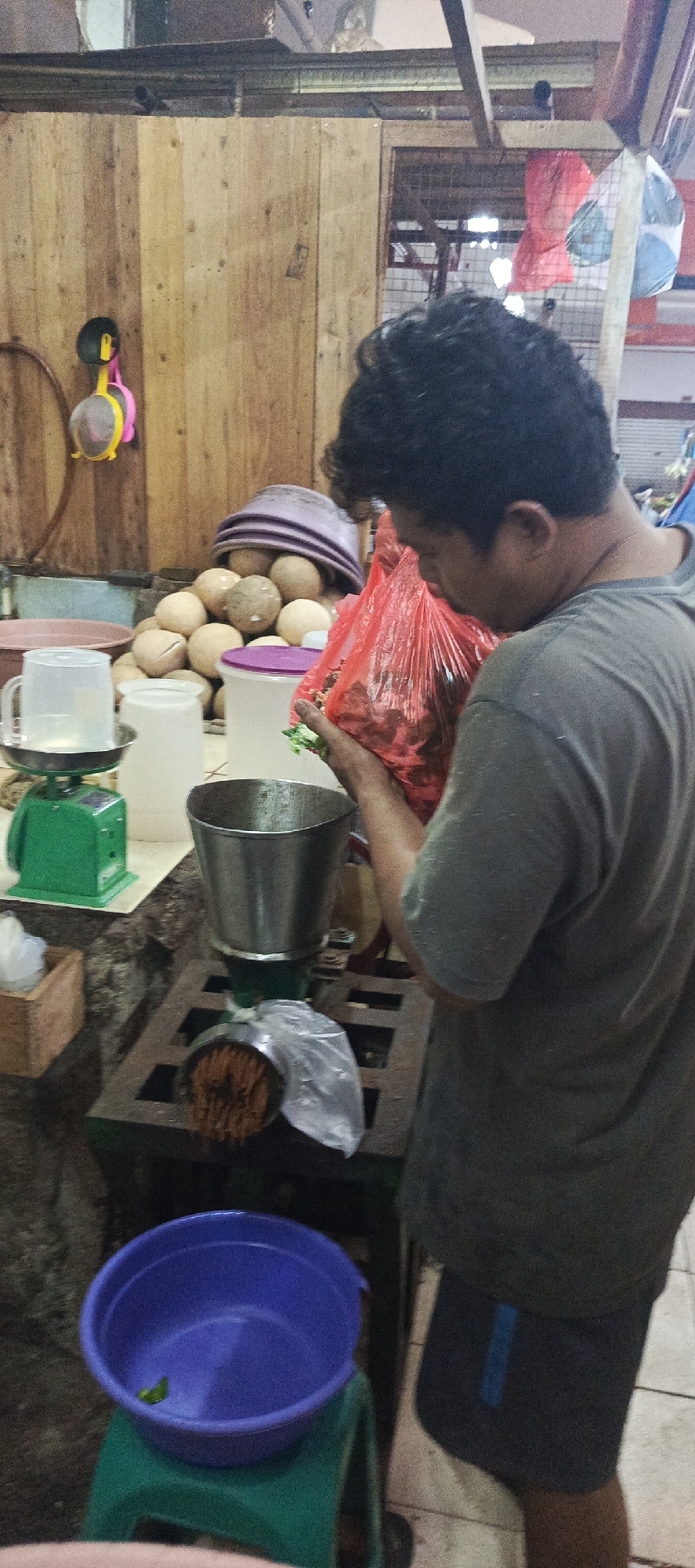 A man in a grey t-shirt and dark shorts is standing in a market in Sukaresmi, Indonesia. He is holding a red plastic bag full of what appears to be red chilies. In front of him is a metal grinder with a stainless steel hopper, and a clear plastic bag collecting the ground material. To the left of the grinder is a green digital scale with a clear plastic jug on it. Behind the scale are several white and pink plastic containers, and a pile of coconuts topped with purple bowls. To the right of the grinder, a large blue bowl sits on a green stool. The background consists of wooden planks and some shelving with various items. The lighting suggests an indoor setting, possibly a market stall.