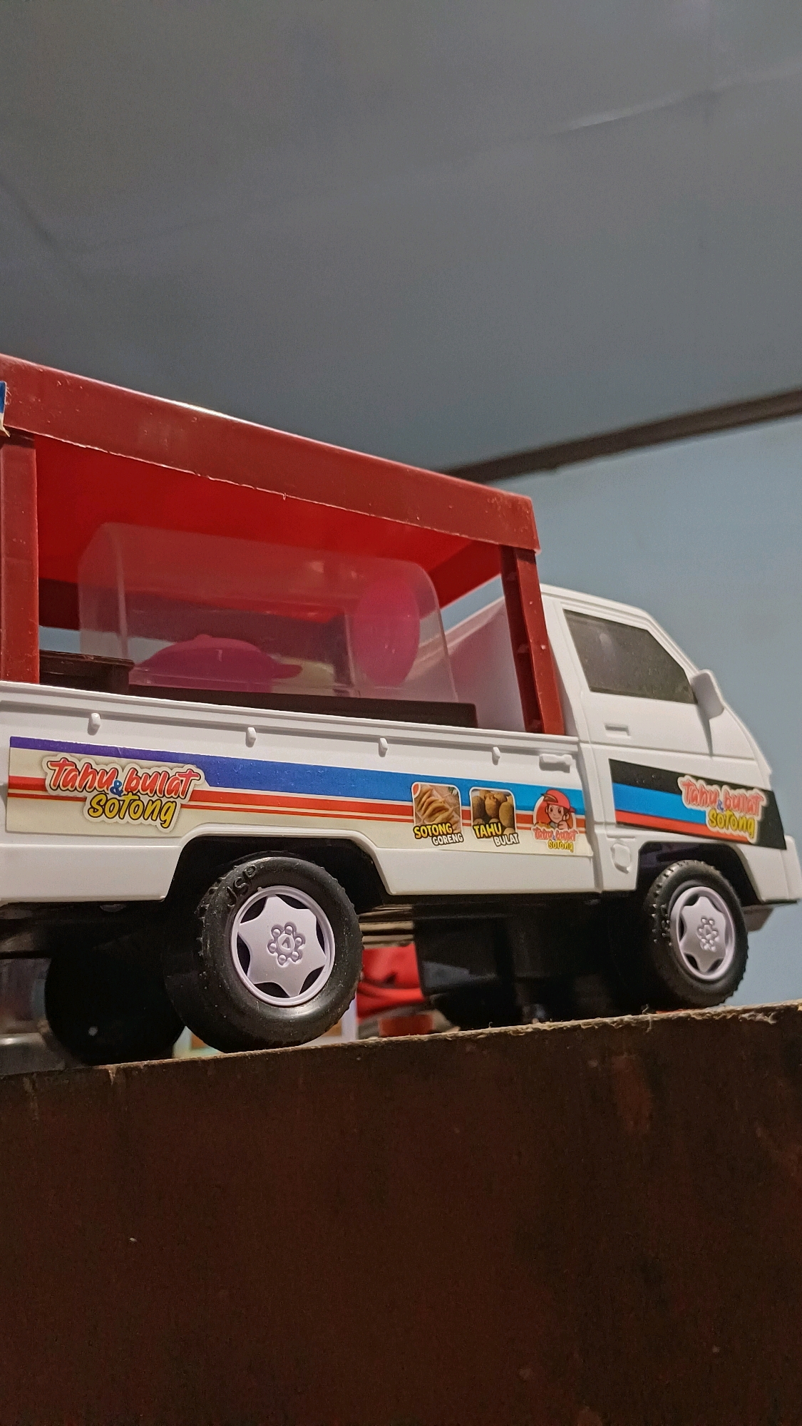 This is a low-angle, close-up, eye-level shot of a white toy pickup truck with a red canopy, viewed from the side. The truck is positioned on a brown, textured surface that appears to be a shelf or a piece of furniture. The background consists of a gradient of gray to light blue. The truck features decals with blue, red, and yellow stripes, and text that reads "Tahu Bulat Sorong." There are also small images of food items labeled "Sotong Goreng" and "Tahu Bulat." Inside the truck's canopy, a pink plastic container is visible. The truck has four black tires with silver hubcaps. The overall lighting is soft, with the scene appearing to be indoors. There are no people visible in the image. The specific location of Wonokerso, Indonesia, cannot be determined from the visual information provided.