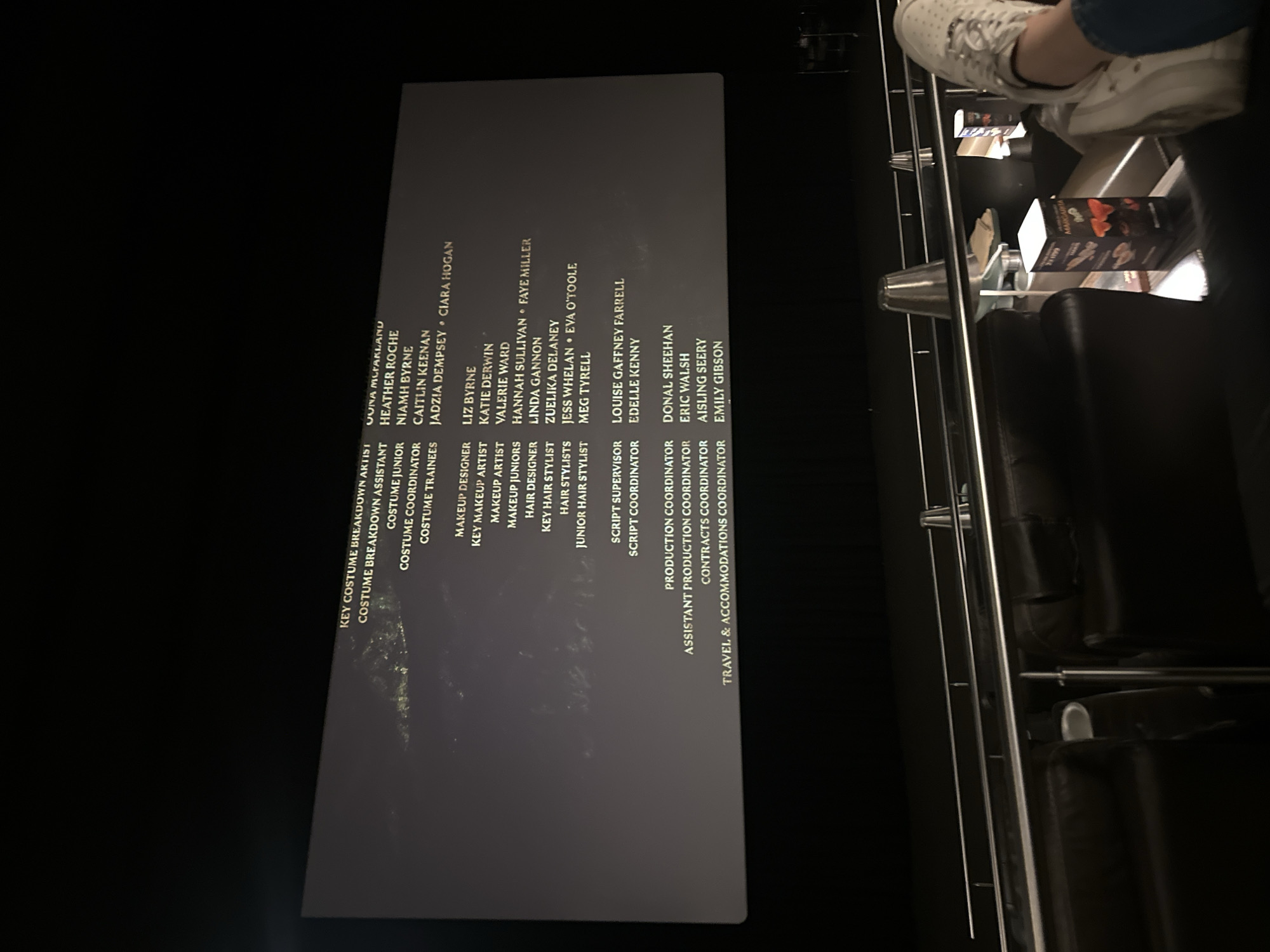 A photograph taken inside a cinema, likely in Huixquilucan de Degollado, Mexico (though this can't be definitively confirmed from the image alone), shows a partial view of a movie screen displaying the end credits of a film. Listed on the screen are various crew member titles and names, including roles in costume, makeup, hair, and production. The camera angle is somewhat tilted. In the foreground, there are black leather cinema seats and a small metal table, upon which rests some snacks. In the upper-right corner of the image, a person wearing white shoes is partially visible.