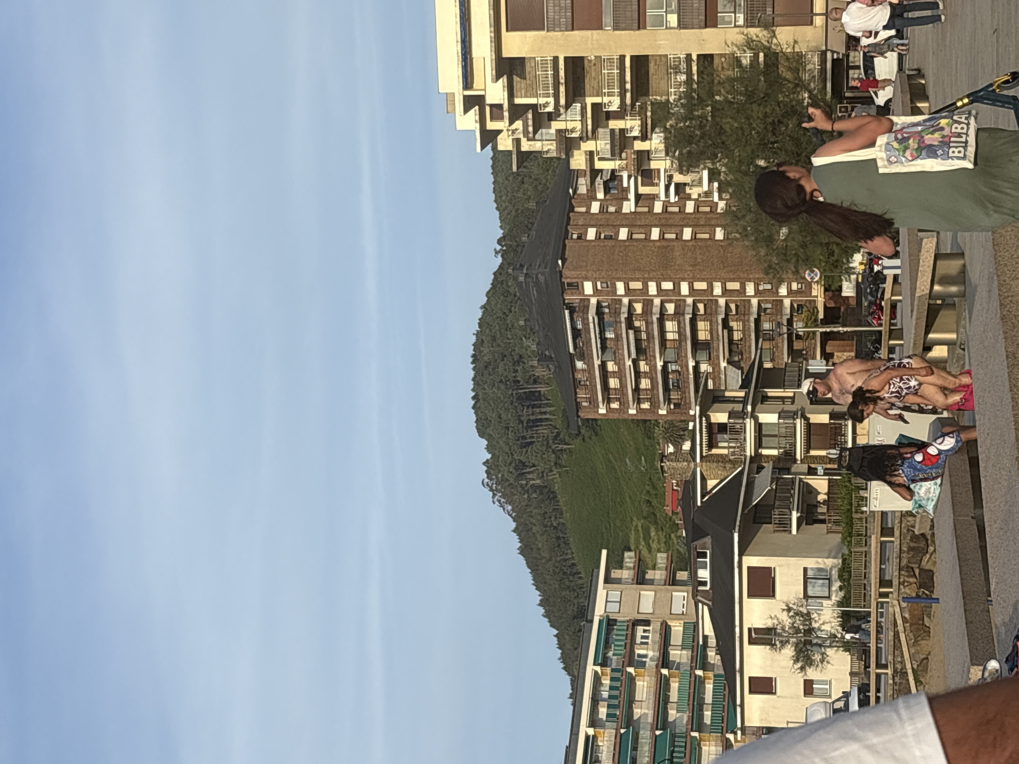 The image shows a street scene in Bakio, Spain, with buildings and people. The sky is clear and blue. In the background, a green, tree-covered hillside rises up. Several multi-story buildings are visible, with balconies and awnings. In the foreground, a group of people are gathered. One person, wearing a green dress and carrying a tote bag, is facing away from the camera. Another person, wearing a white hat and swimming trunks, is also visible. There is a scooter parked on the ground.