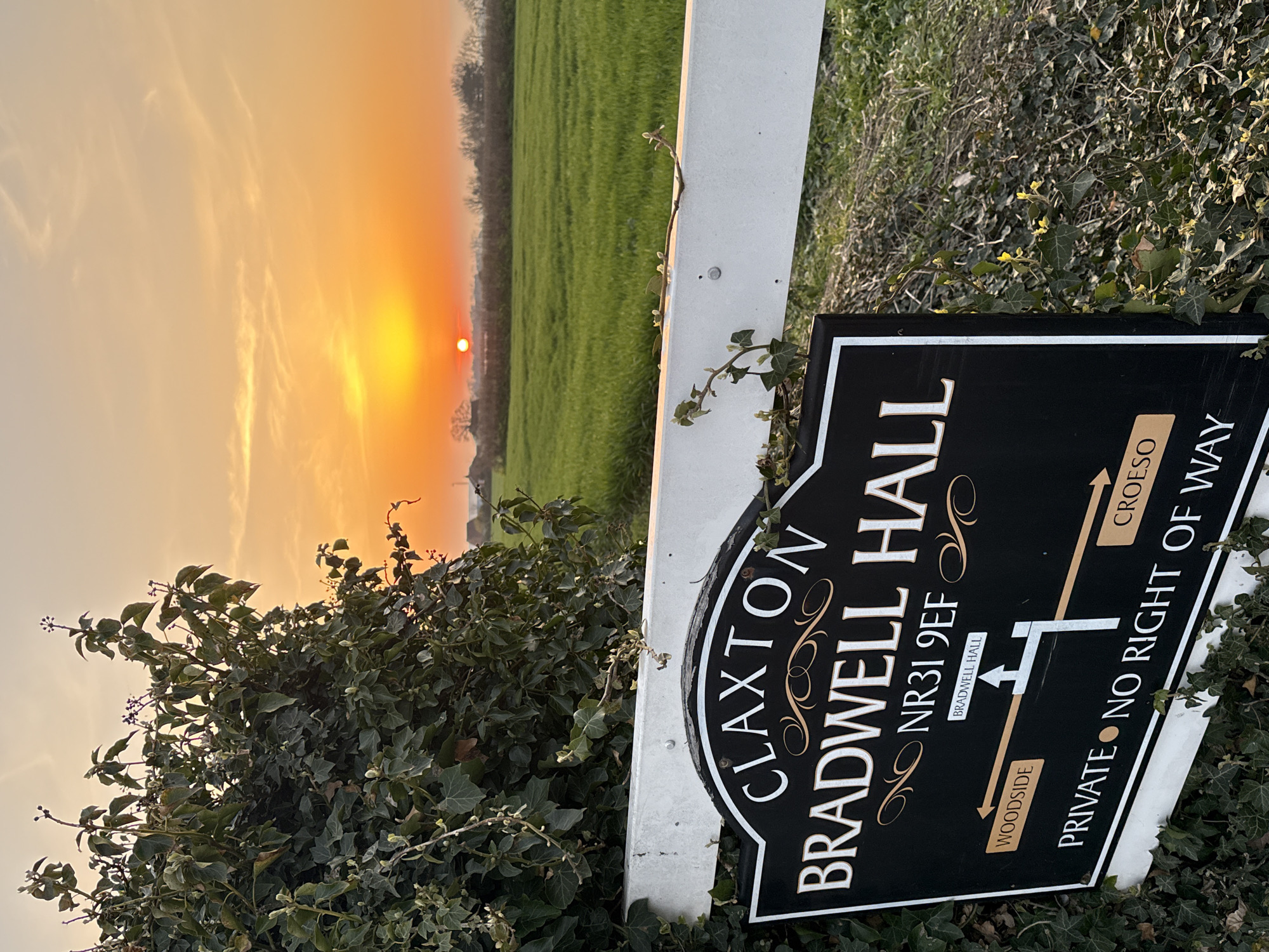 The image is a photograph taken outdoors at dusk or dawn, indicated by the low sun on the horizon and warm sky. The foreground features a signpost with a black sign reading "CLAXTON BRADWELL HALL NR31 9EF" in white and gold lettering, indicating the location. The sign also includes directional arrows indicating "WOODSIDE" and "BRADWELL HALL," and the Welsh word "CROESO" (welcome). A warning, "PRIVATE NO RIGHT OF WAY," is prominently displayed. The signpost is partially obscured by foliage, including ivy. In the background, a green field is visible under the sky. The overall scene appears to be a rural setting near a private residence or estate. The NR31 postcode places the location near the city of Gorleston-on-Sea, United Kingdom.