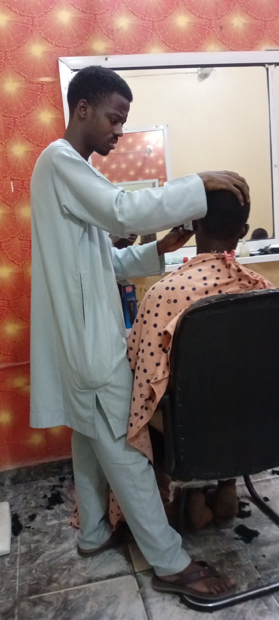 In a room with patterned wallpaper and a tiled floor, a man in a light blue robe and pants is giving a haircut to a person seated in a black chair. The person receiving the haircut is covered with a peach-colored cape adorned with black polka dots. Hair clippings are visible on the floor around the chair. There is a mirror on the wall behind them. The location is Unknown, Unknown.