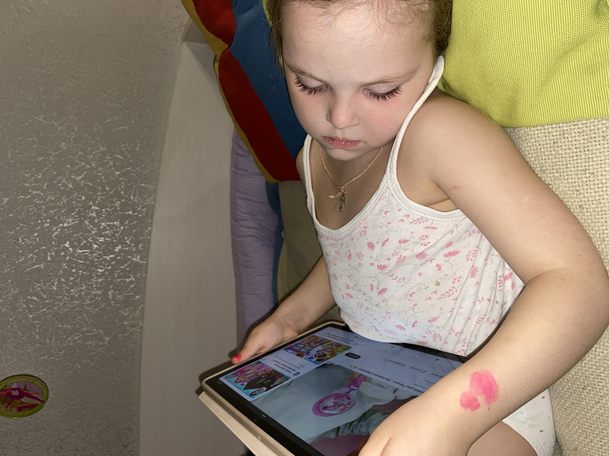 An overhead medium shot captures a young girl, likely between 4 and 7 years old, lying down and engrossed in a tablet device. She has light hair, long eyelashes, and a focused, serious expression as she looks at the screen. She is wearing a white tank top with subtle pink floral patterns and a small cross necklace. A bright pink mark, possibly a temporary tattoo or paint, is visible on her left forearm. Her fingernails appear to be painted red.
The girl is positioned against a light green textured pillow and a beige, woven fabric, suggesting she is on a sofa or bed. Behind her head, a colorful striped fabric in red, yellow, and blue is partially visible. The background features a textured light-colored wall. A small, round sticker with an animated character is affixed to the wall on the lower left.
The tablet screen displays what appears to be a video platform, likely YouTube, showing various video thumbnails. Clearly visible text on the screen includes "5-MINUTE CRAFTS" and "Vidéo pour les parents : Top astuces et découverts!" (French for "Video for parents: Top tips and discoveries!"). The lighting in the room is dim, suggesting an indoor evening or nighttime setting. The scene depicts a quiet, absorbed moment of screen time indoors in Votkinsk, Russia.