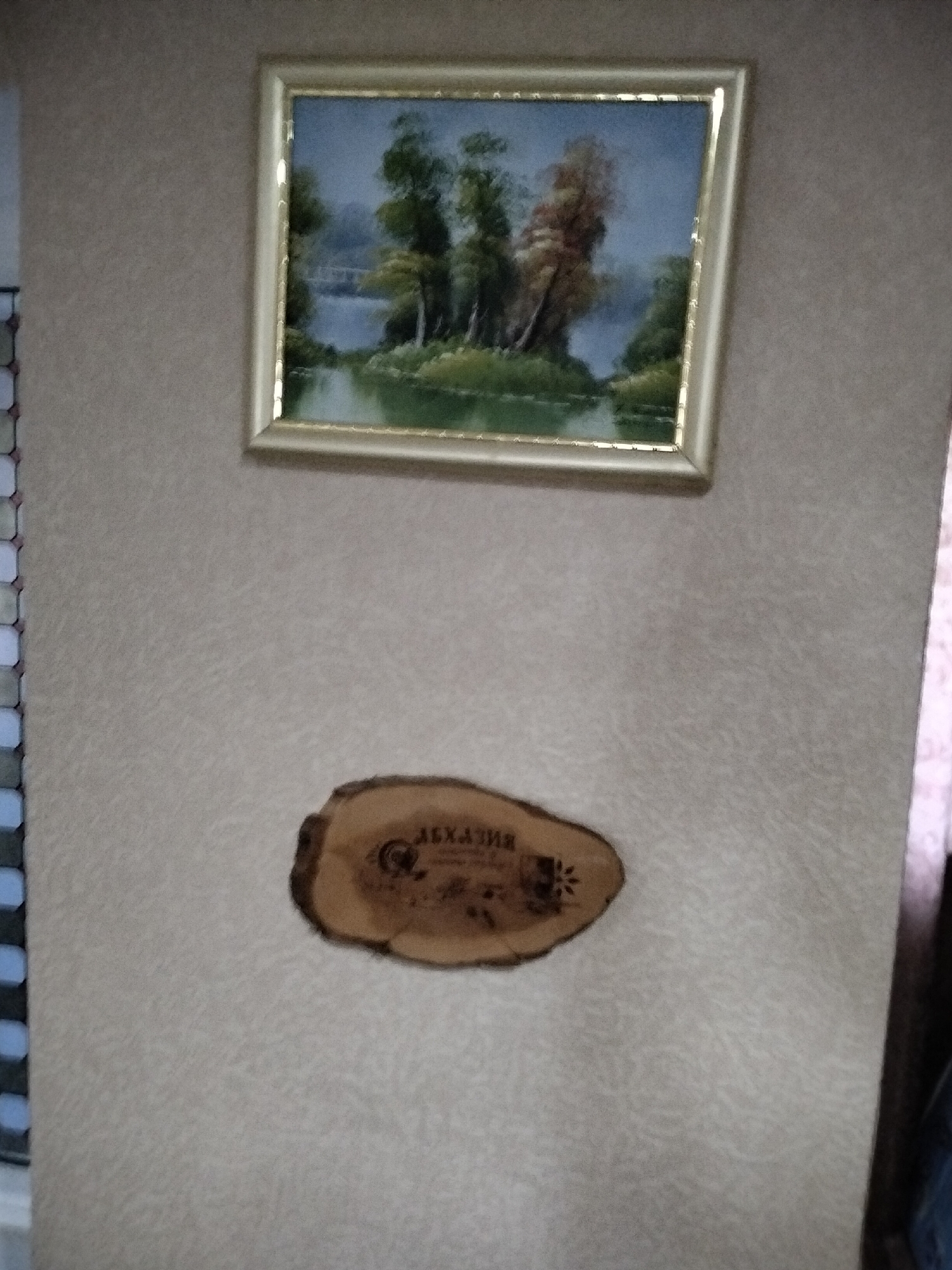 A framed painting of a landscape with trees and water hangs on a textured, light-colored wall. Below the painting, a slice of wood with a burned-in design and text is mounted on the wall. The text on the wood appears to be in Cyrillic script and reads "ЛЕХАЗИЯ". A blurred dark object, possibly a curtain or clothing, is visible on the right side of the image.