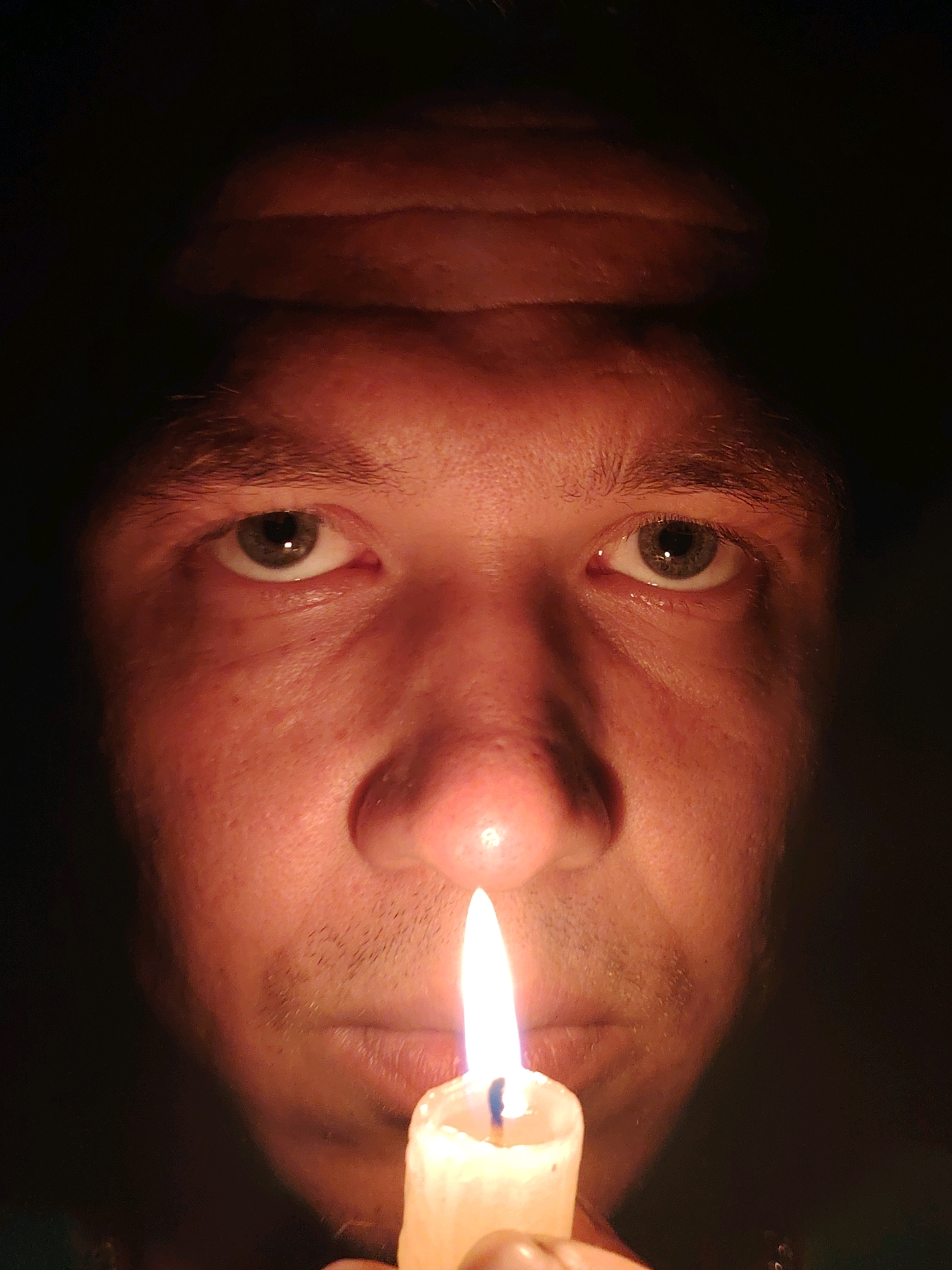 The image is a close-up portrait of a person's face, illuminated by a lit candle held just below their chin. The subject's eyes are open and looking directly at the camera. The lighting creates strong shadows, particularly on the forehead and the sides of the face, highlighting the texture of the skin and the contours of the features. The background is dark and indistinct, focusing all attention on the face and the candle's flame. There is no discernible connection to the city of Chaikovsky, Russia, or any urban environment in this image.