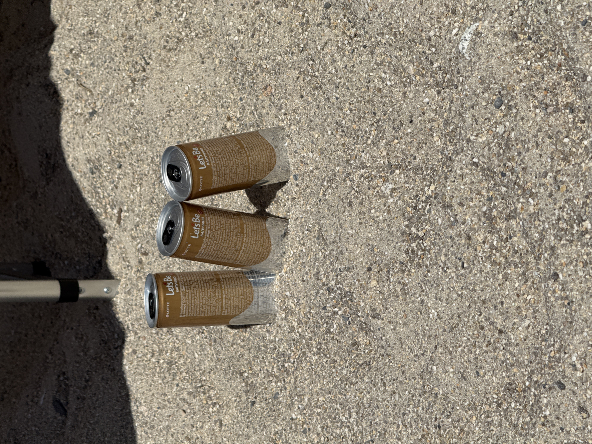 Three cans of "Lets Be Cappuccino" coffee drink, manufactured by LOTTE, are partially buried in sandy soil. The cans are gold-colored with black lettering and logos. They are arranged in a triangular formation. The background consists of dark shadows and what appears to be the leg of a metal structure, possibly part of a table or tent. The soil is composed of small pebbles and sand. The lighting suggests an outdoor setting with a strong light source casting shadows. There is no visible evidence of human presence or activity beyond the placement of the cans. This scene could be located anywhere with such soil and lighting conditions, including the city of Nakhodka, Russia.