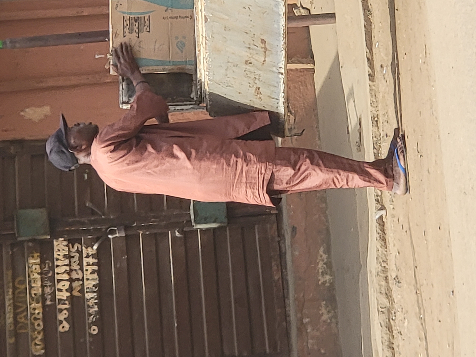An outdoor, brightly lit scene in Maiburiji, Nigeria, depicts a man standing next to a vertical structure, interacting with a cardboard box. The man is positioned on the right side of the image, facing to his left (upwards if the image were correctly oriented). He wears a reddish-brown long-sleeved garment, a dark cap, and blue flip-flops. His hands are placed on an old cardboard box that appears to be taped or affixed to a light-colored, possibly painted, wooden or metal surface, which itself is attached to a larger, darker brown vertical support.
The ground is dusty and sandy, suggesting an unpaved area or a dry environment. Behind the man is a dark brown, corrugated metal gate or door, characteristic of a shop front or a residential entrance. The lighting is harsh and direct, indicating a sunny daytime, likely mid-morning or afternoon.
Visible text includes:
- On the cardboard box: "Creating Better Life" (partially legible) and "10".
- On the metal gate: "DAVIDO", "08140191494", "MAKUS" (or similar, possibly "NICKUS"), "AKUR", and "6 SHARES". These appear to be names and a phone number, likely contact information or advertisements.
The man's gaze is directed towards the box or slightly upwards, but his expression is not visible. He appears to be either attaching something, inspecting, or perhaps retrieving an item from the box.