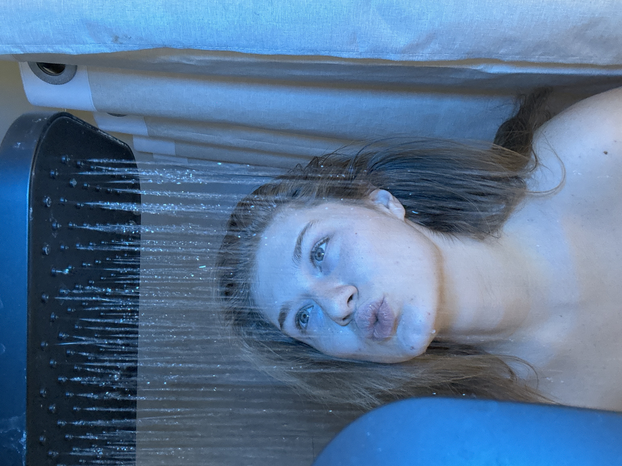 The image portrays a young adult female showering indoors under a dark, rectangular overhead showerhead, emitting multiple streams of water. The entire scene is bathed in blue light. The female, with wet, light brown hair, is shown from the chest up, her shoulders bare. She gazes upwards and slightly to her right, with her lips pursed. Water droplets are visible on her face, neck, and hair. A light-colored fabric shower curtain with visible metal grommets is partially open on the right side of the frame, revealing part of the shower enclosure. A blurred blue object occupies the lower left corner. The environment is a generic shower enclosure, and no specific visual identifiers are present to determine its precise location within Kazan, Russia.