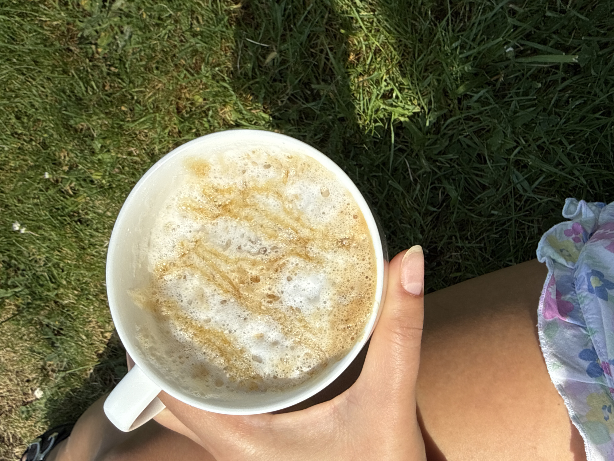 The image shows a close-up, overhead view of a white coffee mug containing a latte with a foamy surface decorated with caramel drizzle. The mug is held by a light-skinned hand. A small portion of a light-colored floral dress is visible on the right side of the frame. The background consists of green grass with varying light conditions, indicating outdoor natural light. The location is unspecified, but based on the prompt, it is assumed to be somewhere in the city of Leicester, United Kingdom. No people or actions beyond holding the mug are visible.