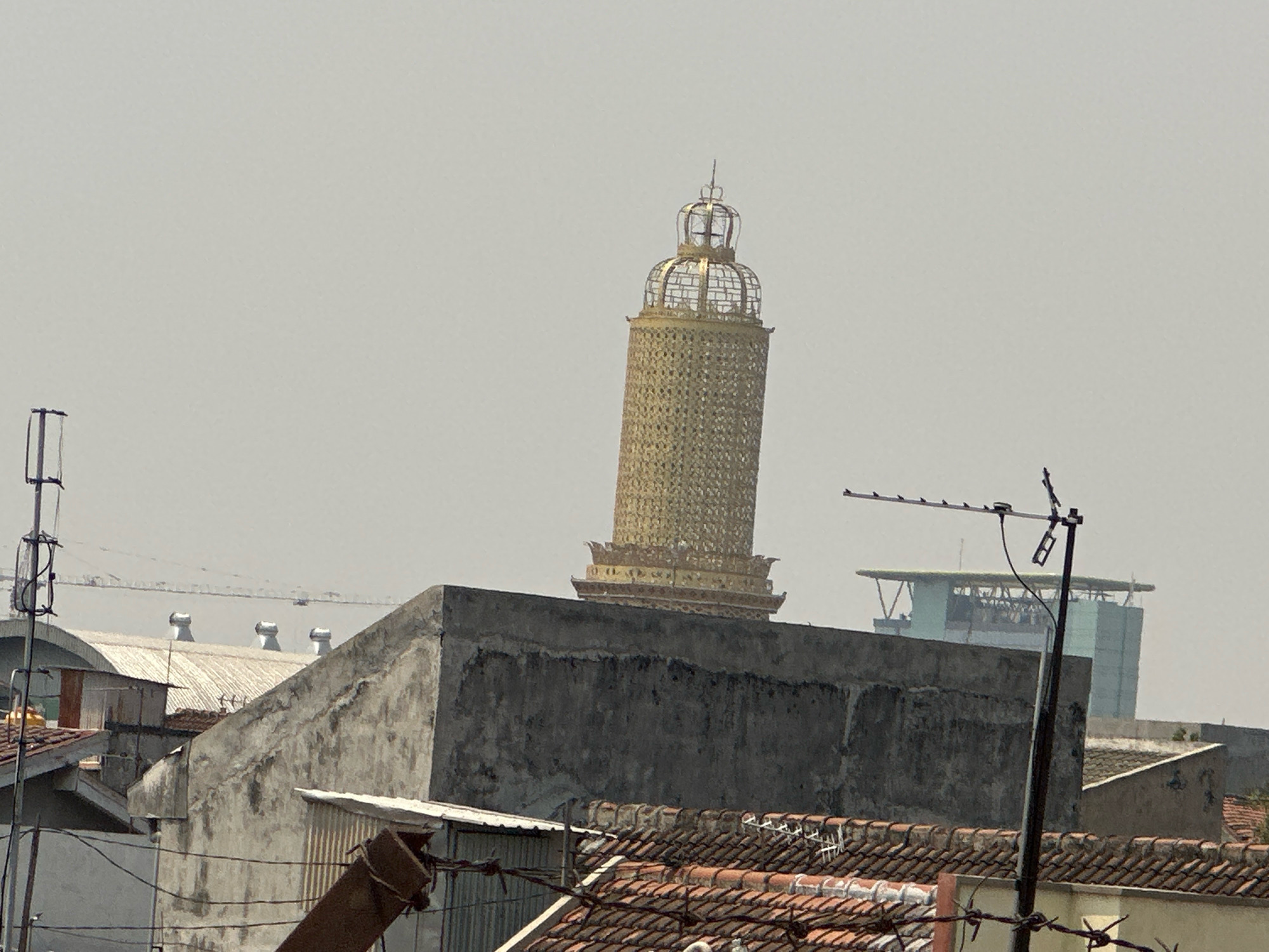 The image depicts a cityscape with a prominent golden tower-like structure in the background. The tower is cylindrical and appears to be made of a metallic material, possibly gold-plated, with intricate detailing and a cage-like crown at the top. In the foreground, there are rooftops of buildings, including some with tiled roofs and others with corrugated metal. A concrete wall partially obstructs the view of the tower. Antennas and power lines are visible, suggesting an urban environment. In the far background, other buildings and structures can be seen, some of which are modern in design with glass facades. The sky is overcast and a pale grey. The scene is likely an outdoor shot taken during the daytime, as indicated by the bright lighting. This location is identified as Purwotengah, Indonesia.