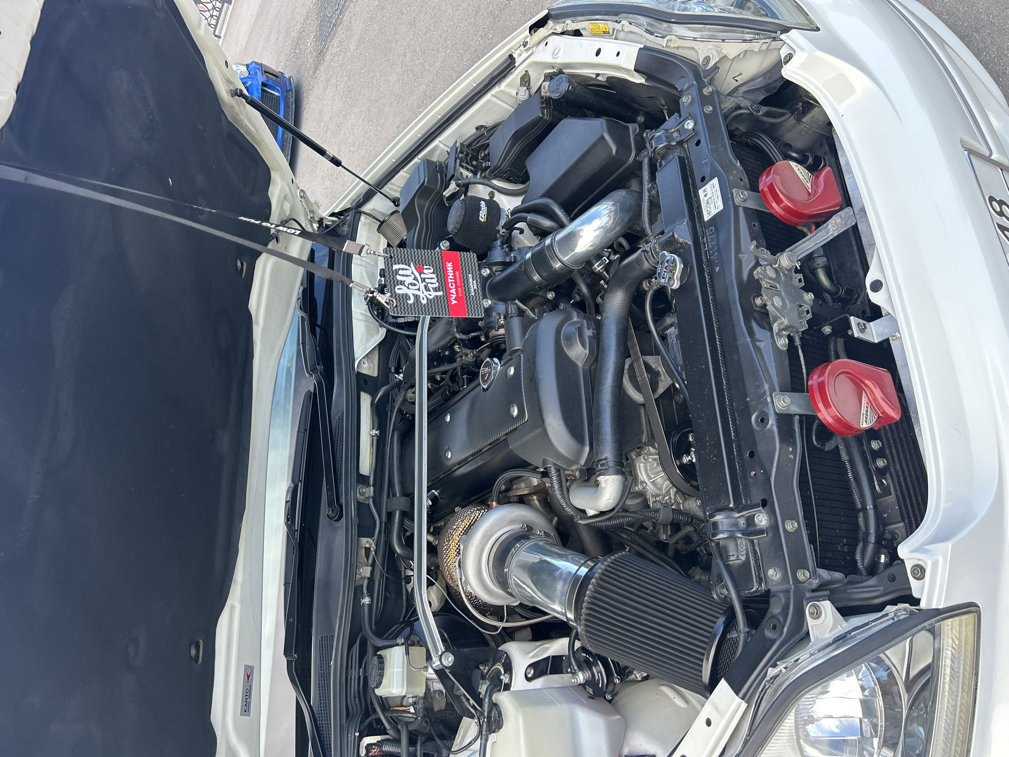 The image shows the open engine bay of a white car. The engine compartment is filled with various components, including a large turbocharger, air intake system, and engine block. A black lanyard with a tag hangs from the hood support, displaying "Low Fun" and "УЧАСТНИК" (Participant) in Russian, along with "Khabarovsk 2024". Two red aftermarket horns are visible at the front of the engine bay. In the background, a blue car is parked on an asphalt surface. The setting appears to be an outdoor event or car show in Khabarovsk, Russia, likely during daylight. No people are visible in the frame.