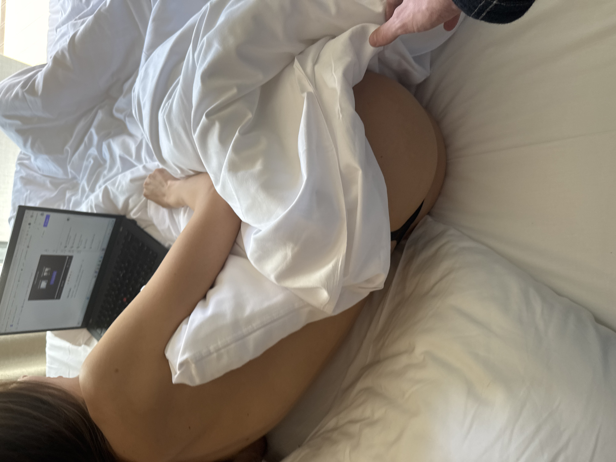 The image presents a top-down view of a person, likely a woman with long dark hair, lying on a bed covered in white sheets and pillows. She is partially nude, with her bare back and arm visible, and appears to be wearing black underwear. A black laptop is positioned next to her, displaying what looks like a web browser with a video playing and other interface elements, though specific text is unreadable.
In the upper right corner of the frame, another person's hand, with visible wrist hair and a dark fabric sleeve, is gently pulling back the white duvet from the first person's lower body, revealing her buttocks and the underwear. This action suggests an intimate or playful moment. The bed linen is slightly rumpled, indicating recent use. Soft, natural light illuminates the scene, suggesting it is daytime, possibly morning, filtering in from a source off to the left. The setting is clearly an indoor bedroom. The interaction between the two individuals is central to the image, conveying a sense of casual intimacy and warmth.