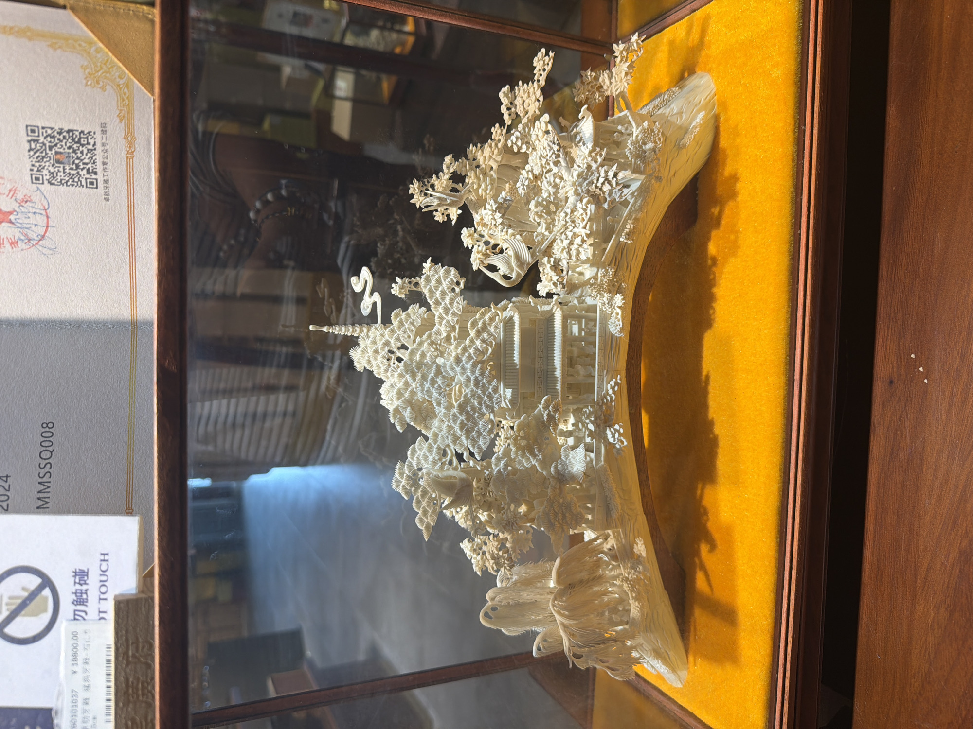 An exquisitely intricate ivory carving, likely Chinese in origin, is displayed within a wooden and glass case with a vibrant yellow velvet backing. The carving itself depicts a detailed miniature landscape featuring traditional pagodas, temples, and dwellings nestled among dense, finely sculpted trees and foliage. Subtle reflections on the glass surface show hints of the surrounding room, including what appears to be a person's arm wearing a beaded bracelet.
To the left of the carving, a certificate or informational placard is visible, partially obscured. The text on the placard includes "2024," "MMSSQ008," and a price of "¥ 18800.00." A prominent warning "勿触碰" (DO NOT TOUCH) is also visible, with an incomplete English translation "OT TOUCH." Further text identifies the source as "卓航牙雕工作室公众号二维码" (Zhuohang Ivory Carving Studio Official Account QR Code), confirming the material as ivory. A QR code and a red stamp are also present on the placard. The artwork is presented as an exhibit, possibly in a gallery, museum, or specialty shop, emphasizing its value and delicate nature.