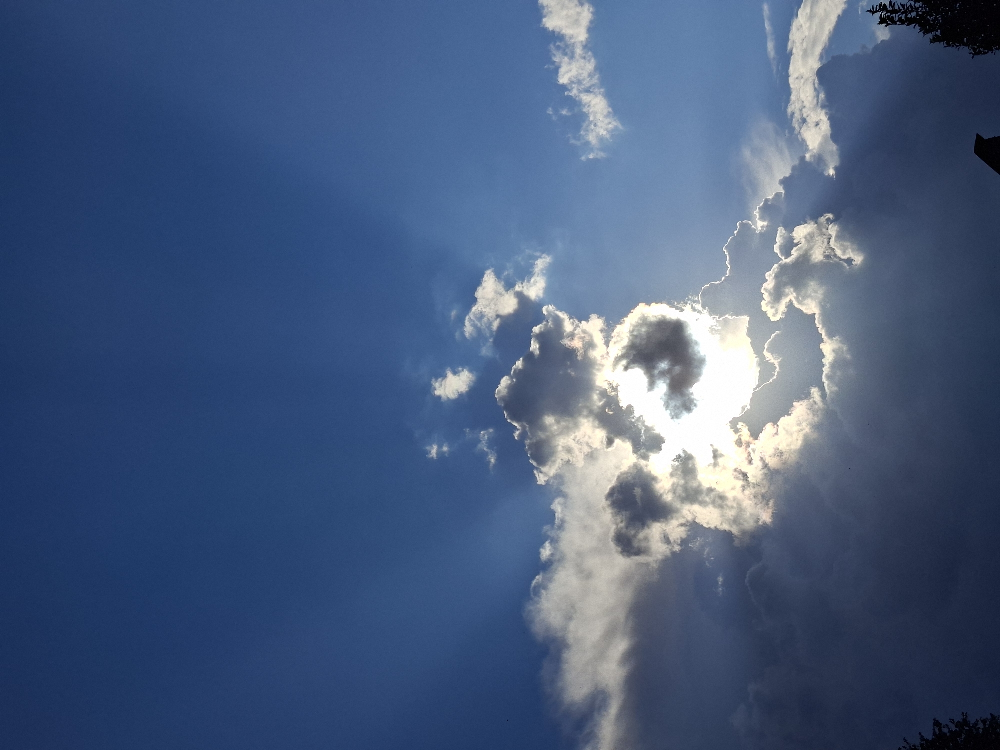 The image shows a dramatic sky with sunlight breaking through dark clouds. The sky is a deep blue, and rays of light emanate from behind a cluster of clouds in the center of the frame. Some clouds are bright white and illuminated by the sun, while others are dark and shadowed. In the lower right corner, a silhouetted tree and part of a roof are visible against the sky. The overall scene evokes a sense of either an approaching storm or the aftermath of one, with the sun piercing through the remaining darkness.