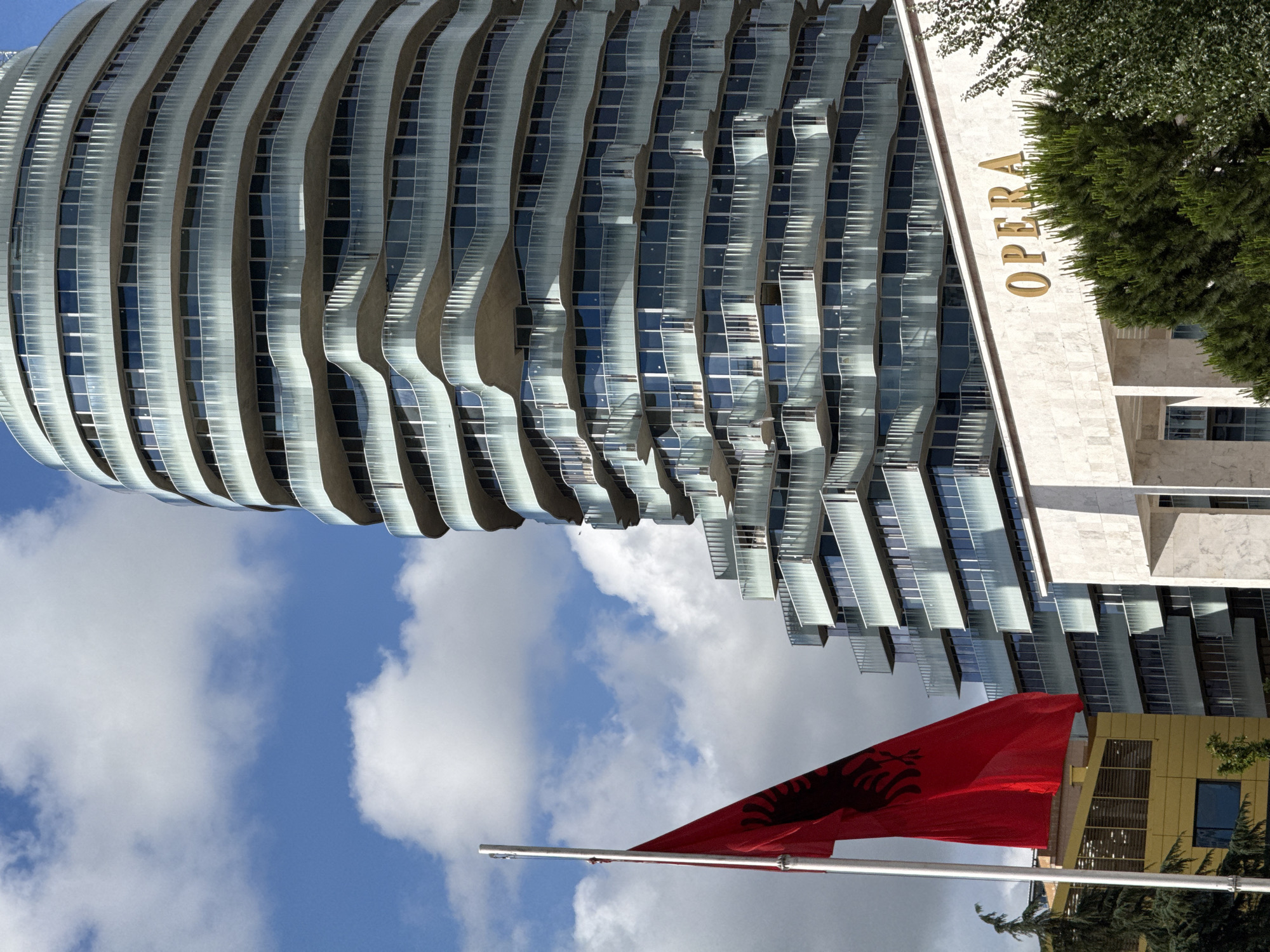 This media file captures a vibrant urban scene in Tirana, Albania, on a bright, partly cloudy day. The dominant feature on the left is a tall, modern skyscraper characterized by its distinctive wavy and layered architectural design. Its facade is composed of numerous curved, reflective glass panels and light-colored horizontal elements, mirroring the blue sky and scattered white clouds.
To the right of this contemporary structure stands a building with a more traditional appearance, possibly clad in light-colored stone. The word "OPERA" is prominently displayed vertically in golden letters on its facade, partially framed by lush green trees and foliage.
In the foreground, at the bottom center of the image, a bright red Albanian flag, emblazoned with the iconic black double-headed eagle, waves from a white flagpole.
The scene showcases a dynamic blend of modern and classic architecture under clear daylight conditions, with no specific human activity visible. The presence of the Albanian flag and the "OPERA" inscription provide strong contextual cues for the location in Tirana.