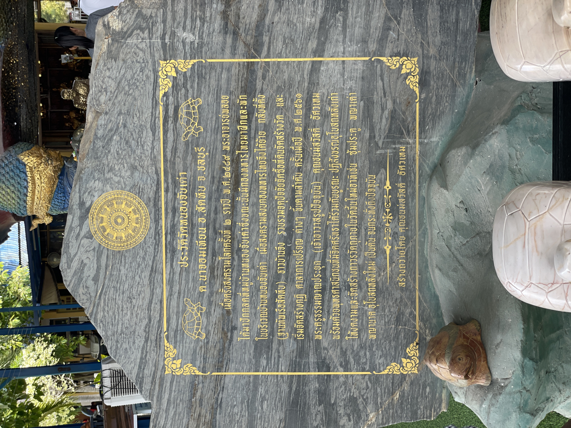 The image shows a large, dark grey stone inscribed with Thai text and golden ornamentation. The text appears to be a historical marker or dedication. At the top center of the stone is a golden emblem, possibly a Buddhist symbol. Two golden turtle outlines are present, one on the left and one on the right, framing the main text. The text describes the history of a temple, mentioning dates and important figures. Below the main text, it states "สร้างถวายโดย คุณชนม์มัสสวัสดิ์ อัศวเขรม" which translates to "Donated by Khun Chonmasat Assawakrom". The stone is situated outdoors, with greenery visible in the background and foreground. To the lower left of the stone, a small, carved brown turtle sits on a green rock. To the lower right, part of a white, carved turtle shell is visible. The overall impression is of a cultural or religious monument in a natural or landscaped setting. The inscription mentions "Tambon Na Chom Thian" and "Chonburi", indicating the location.