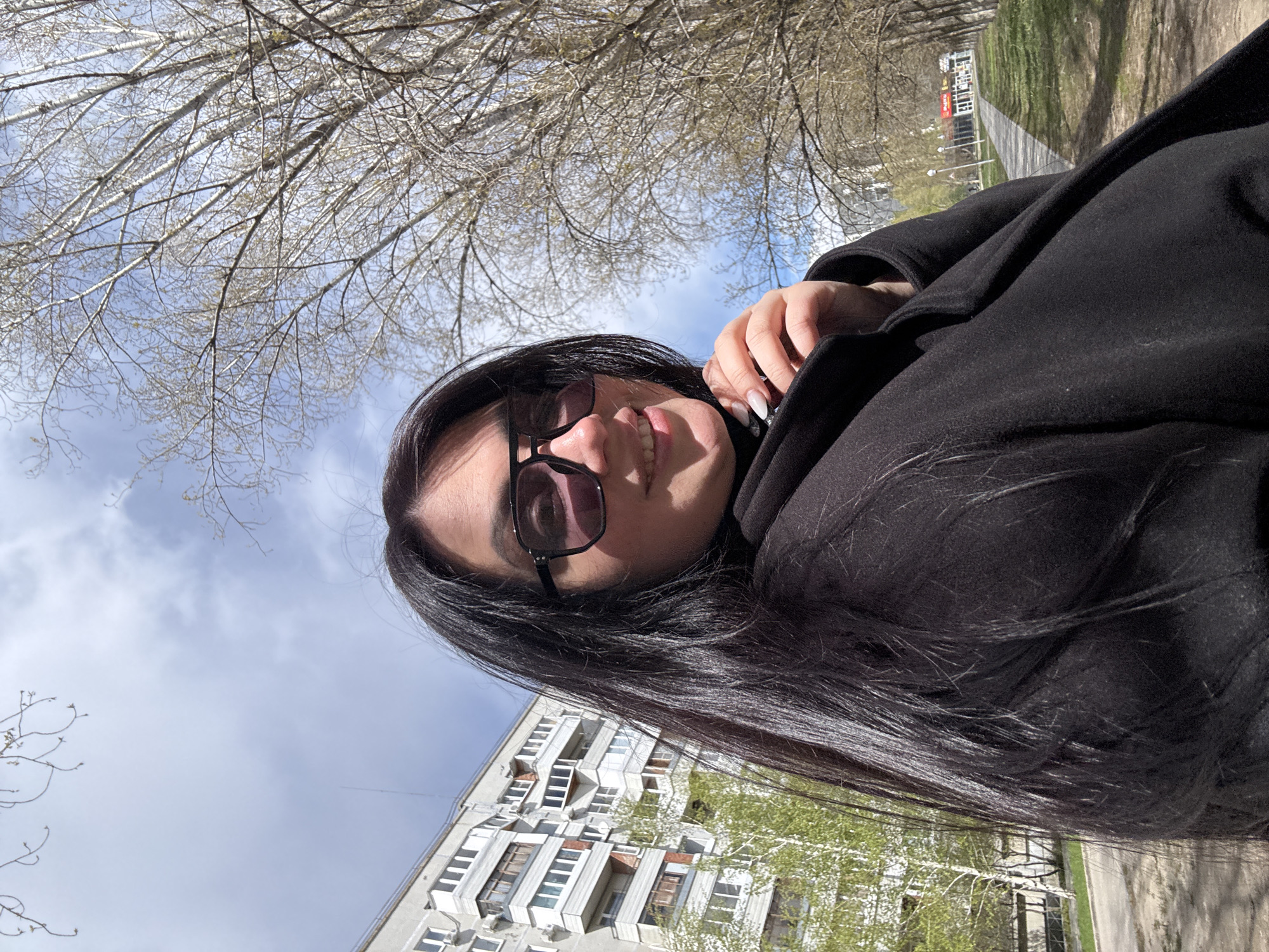 The original image is rotated approximately 90 degrees counter-clockwise. Described from a corrected upright orientation, the photograph is a close-up selfie of a woman outdoors, smiling and looking slightly upwards. She has long, straight dark hair, wears dark-rimmed sunglasses, and a black coat. Her left hand, with long, pointed white fingernails, is visible near her chin or collar.
The setting appears to be a park or green space adjacent to a residential area. Above the woman, numerous bare tree branches are silhouetted against a bright blue sky with a few scattered white clouds, suggesting late winter or early spring. To the left of the frame, a multi-story residential building with a grey facade, featuring multiple windows and balconies, is partially visible. Its architectural style suggests a post-Soviet or Eastern European origin. To the right and further in the background, a paved path, green grass, and more trees are present. In the far distance on the right, a building with a red sign can be seen, but any text on it is unreadable.
The bright lighting indicates it is daytime. The woman's smile conveys a happy or relaxed mood.