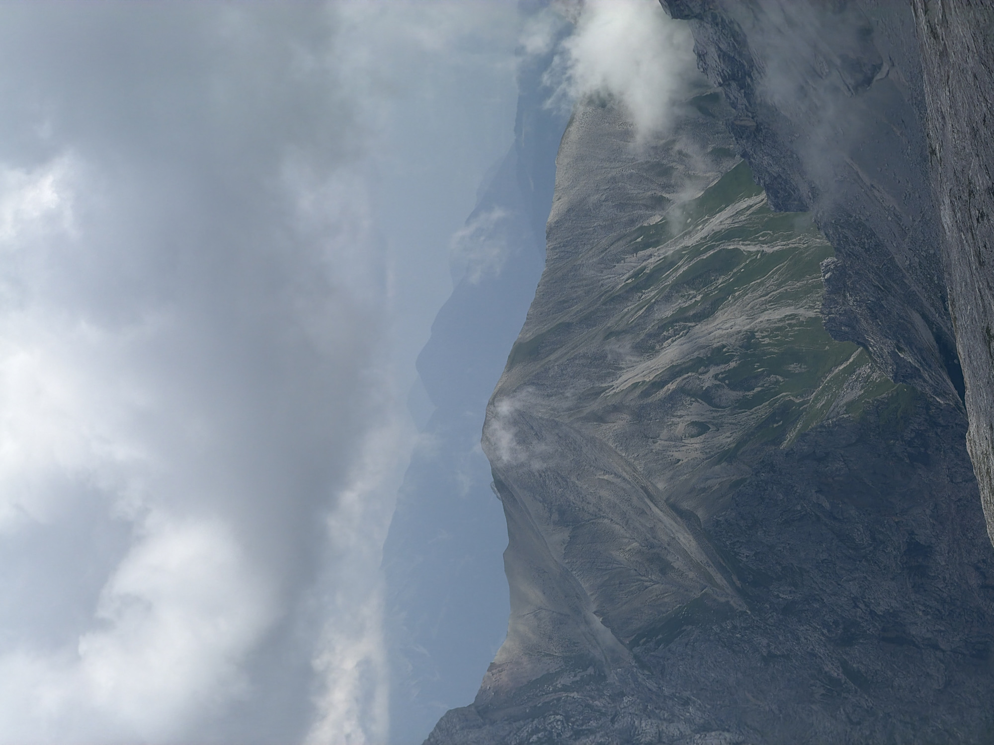The image depicts a dramatic mountain landscape under a cloudy sky. The foreground features rugged, rocky terrain, with sharp ridges and steep slopes. In the mid-ground, a prominent mountain ridge stretches across the frame, its slopes a mix of scree, sparse vegetation, and bare rock. Wisps of white clouds cling to the higher elevations, partially obscuring the mountain peaks and adding to the atmospheric quality of the scene.
Further in the distance, layers of mountains recede into a hazy, blue-toned background, suggesting a vast and expansive mountain range. The sky is filled with heavy, gray clouds, indicating overcast conditions, possibly with a recent or imminent rain shower. The lighting is diffused, casting a soft, even light over the landscape, with no strong shadows.
This scene is characteristic of the Bavarian Alps, and specifically the dramatic peaks found in the vicinity of Garmisch-Partenkirchen, Germany. The ruggedness, scale, and geological features are consistent with the prominent mountains in this region, such as the Zugspitze or the surrounding massif. There are no people or man-made objects visible in the image, focusing solely on the natural, untamed beauty of the alpine environment. The overall impression is one of grandeur, solitude, and the raw power of nature.