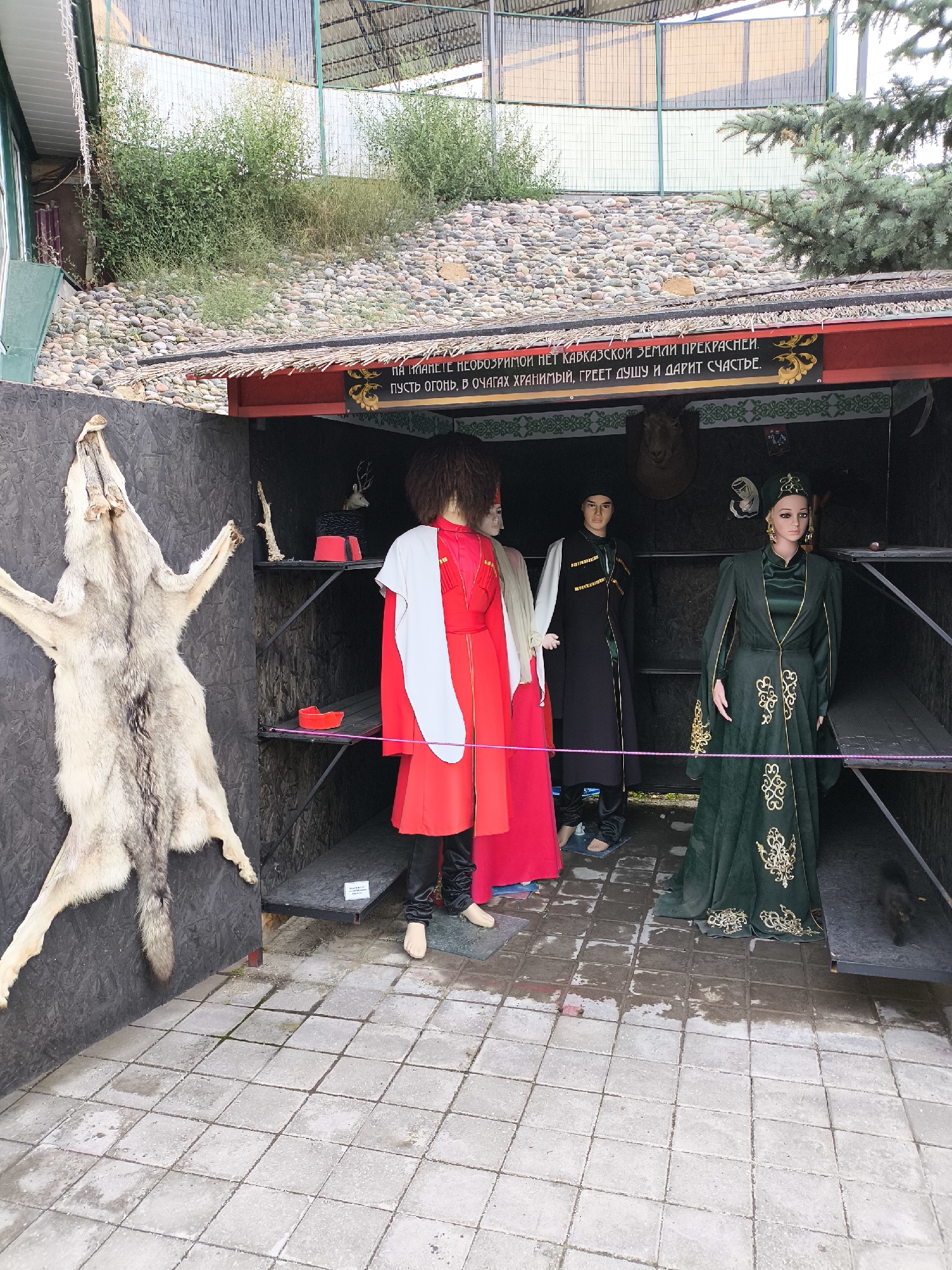 In Borgustanskaya, Russia, an outdoor stall displays several mannequins dressed in traditional attire and animal pelts. To the left, a large wolf pelt is mounted on a dark gray wall. Inside the stall, three mannequins stand on a tiled floor. The first mannequin on the left wears a long red robe with white accents and a large, dark, curly wig. The second mannequin, positioned behind and slightly to the right of the first, wears a black robe with gold trim and a dark head covering. The third mannequin, to the right, is adorned in an elaborate dark green robe with gold embroidery and a matching head covering, and large gold earrings. Behind the mannequins, shelves hold various items, including a red bowl, a taxidermied deer head, and decorative elements. The stall itself is constructed with dark wood and features a thatched roof. The ground in front of the stall is paved with light gray square tiles. The background consists of a stone-covered embankment with green foliage and a sloped roof structure with translucent panels.