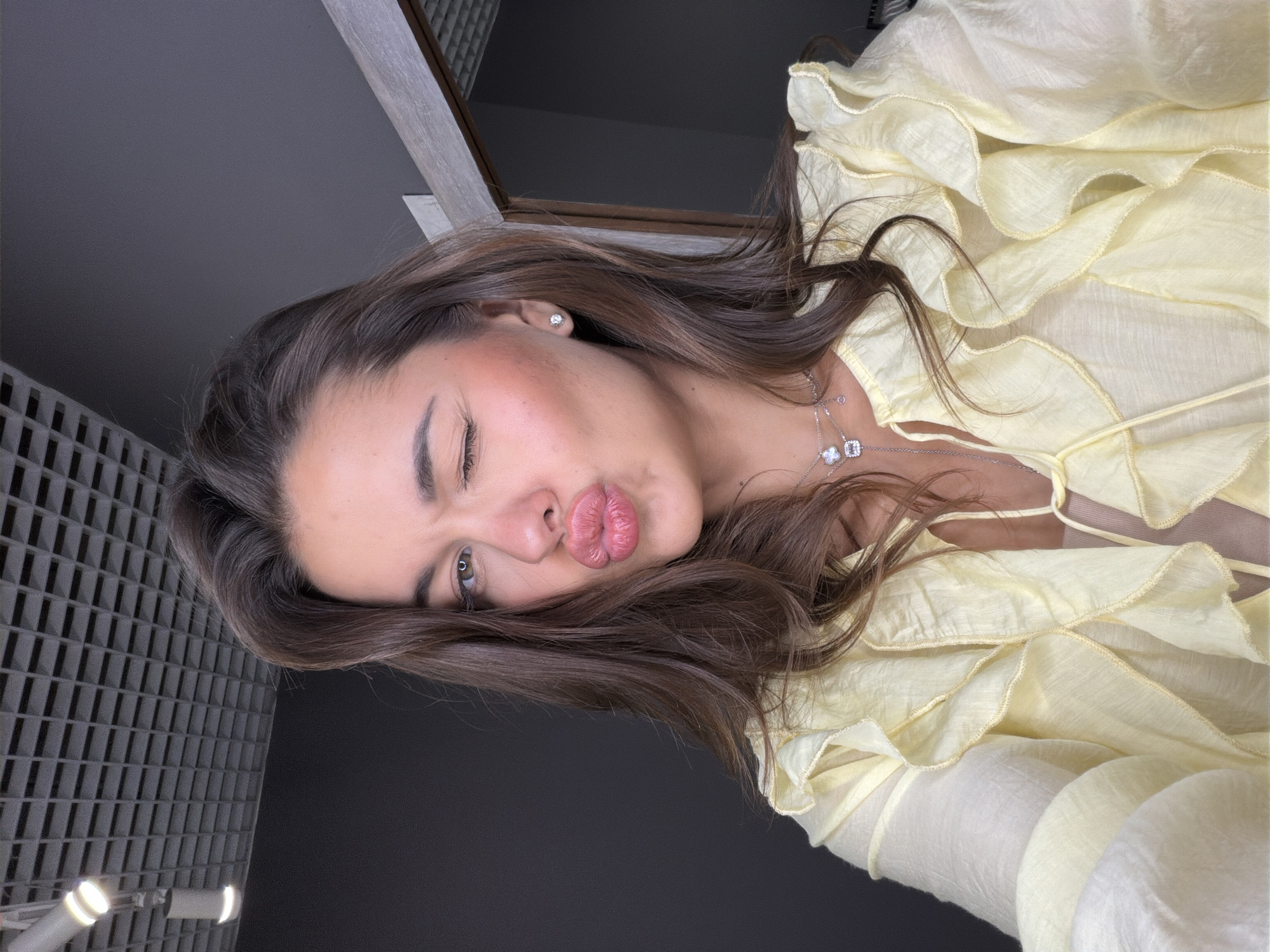 A medium-shot, eye-level, portrait-oriented photograph shows a woman with brown hair and fair skin in an interior location. She is wearing a light yellow, ruffled blouse and a delicate silver necklace. The woman is puckering her lips in a kiss and winking one eye. The background consists of a gray padded wall, possibly soundproofing material, and an edge of a picture frame. The lighting appears to be artificial and illuminates the subject's face. No information is available to determine the location in the city of Krasnoyarsk, Russia.