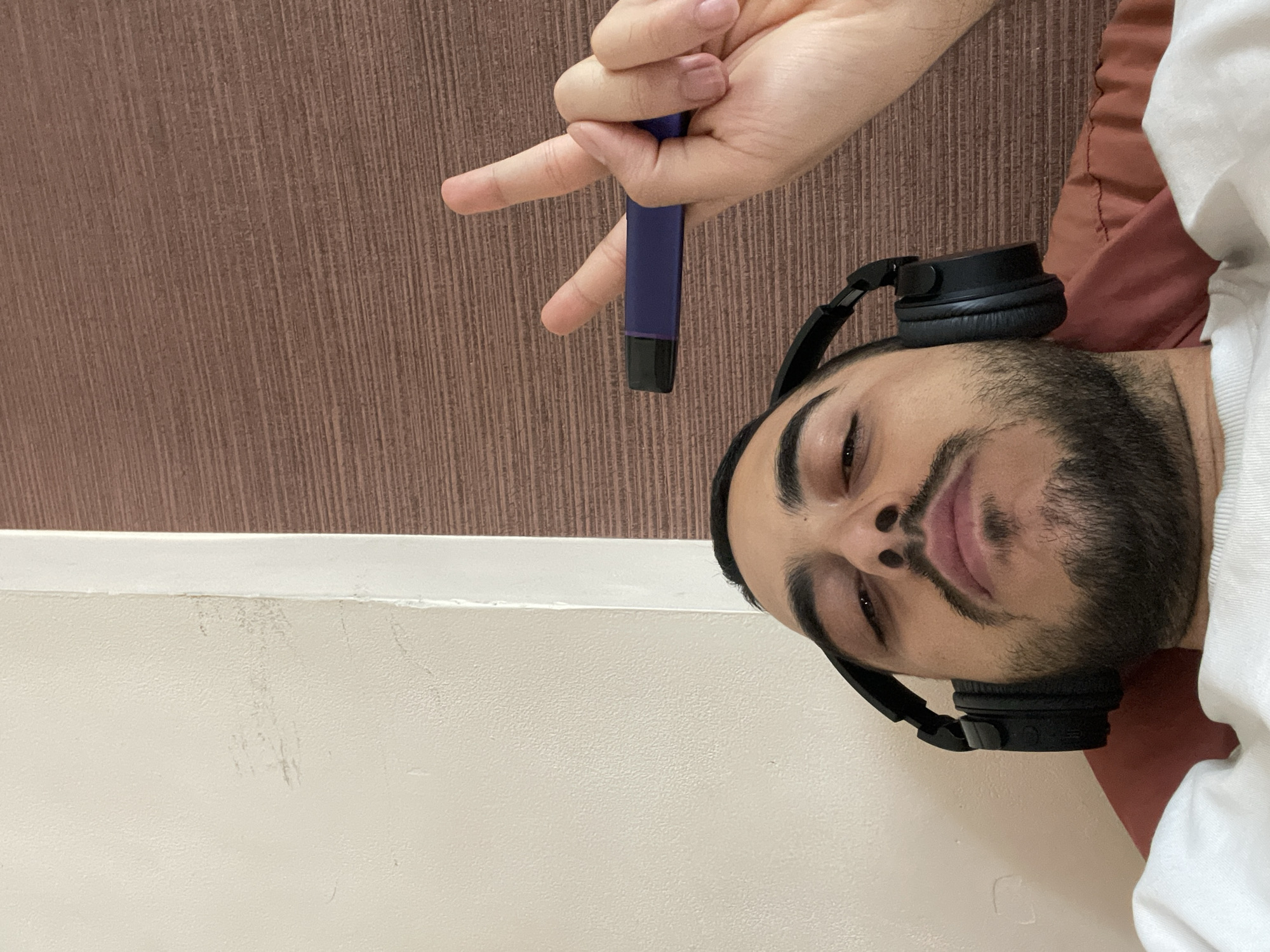 A man lies supine, wearing black headphones, with a purple cylindrical object held above his head by a hand. He is in an interior space with a white wall at the bottom left and a brown wall in the background. A pillow or cushion is visible to his right. The location is unspecified, and Varna, Bulgaria is not evident.