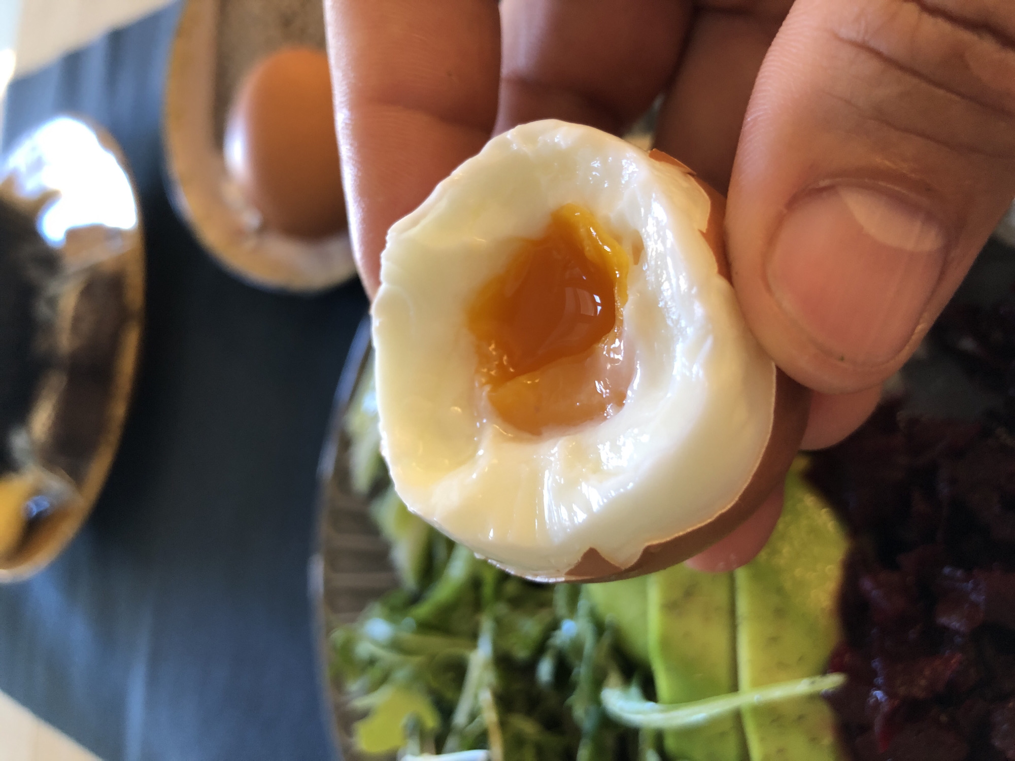 A close-up, high-angle indoor photograph displays a human hand holding a soft-boiled egg, positioned centrally in the frame. The egg is partially peeled, revealing a firm white albumen encircling a distinctly liquid, bright orange yolk that reflects light. The unpeeled portion of the egg shows its brown shell. The hand, primarily the thumb and index finger, exhibits clear skin texture and clean fingernails, gripping the egg from below.
The background is out of focus, presenting components of a meal arranged on a dark, possibly grey or black, surface, which may be a placemat. To the left, a bowl contains green leafy vegetables, resembling arugula, with slices of light green avocado positioned beneath them. Further below the avocado, a dark red or purple granular substance, potentially diced beets, is visible. To the right, a whole, brown-shelled egg rests upright in a small, light-colored egg cup. In the upper background, a blurred ceramic object, possibly a mug or bowl with a handle, reflects bright light from its rim. The overall lighting is bright and even. The image contains no discernible visual cues that would allow for the identification of Saint Petersburg, Russia, as its specific location.