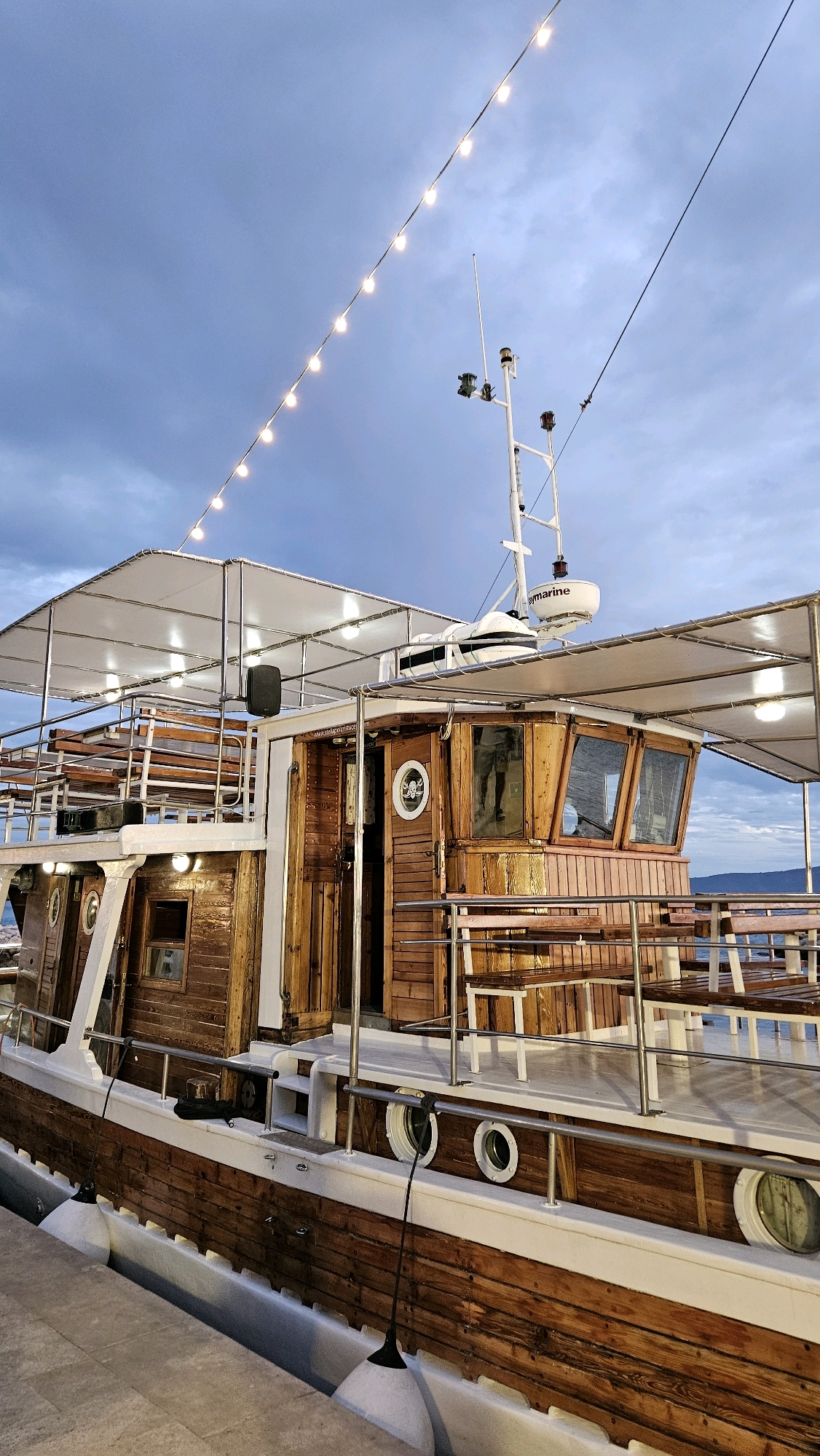 The image depicts a wooden boat docked at a seaside location, likely in Općina Tučepi, Croatia. The boat features multiple decks with wooden benches and a covered upper deck illuminated by string lights. A pilot house with large windows is visible, equipped with marine electronics, including a radar dome. The boat's hull is made of dark wood with white trim, and white fenders are suspended along its side. In the background, the sky is overcast with muted blue tones, and a distant coastline with hills can be seen. The overall scene suggests a calm, dusky atmosphere. There are no people clearly visible in the image, and the focus is on the boat and its surroundings.
