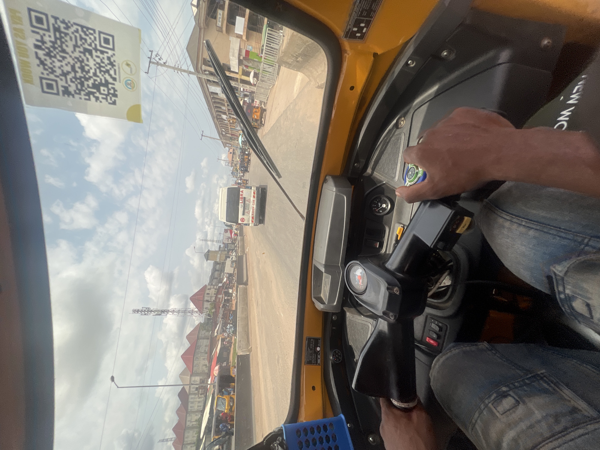 The image is taken from inside a yellow motorized tricycle, commonly known as a "Keke Napep", as it navigates a busy street in Onitsha, Nigeria. The driver's hands are visible, one on the ignition and the other on the controls. The view through the windshield shows a street lined with buildings, some with corrugated iron roofs. A white minibus is approaching. Overhead power lines are visible. A QR code is affixed to the upper left of the windshield. The driver is wearing jeans.