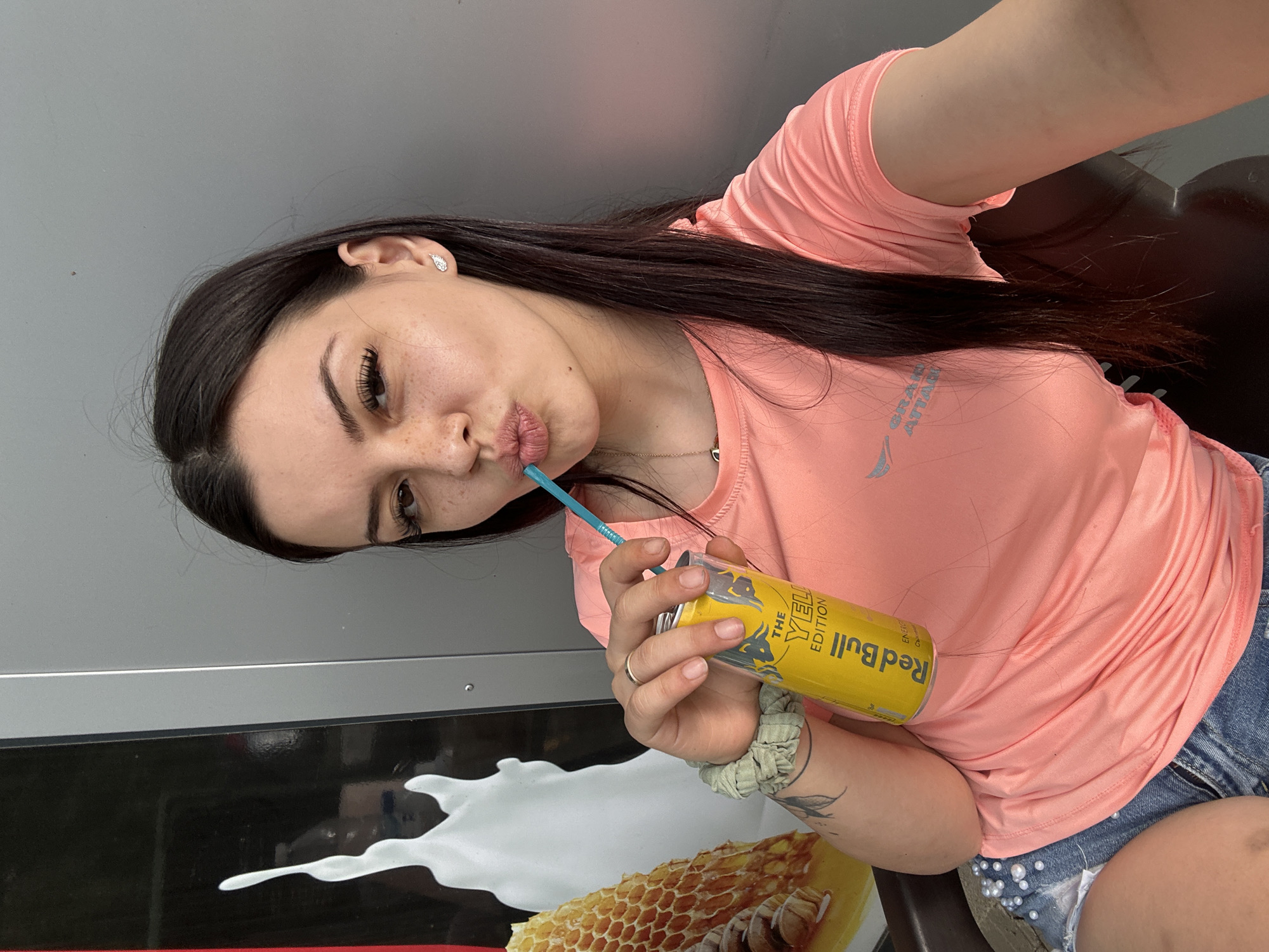 A young woman with dark hair and freckles is posing for a selfie while holding a can of Red Bull with a blue straw in her mouth. She is wearing a pink t-shirt and denim shorts. She has a scrunchie on her left wrist and a ring on her right ring finger. She is indoors, and the background shows a grey wall and a large advertisement with an image of milk and honeycomb. The advertisement appears to be for a dairy product. The woman is looking at the camera and has her lips pursed.