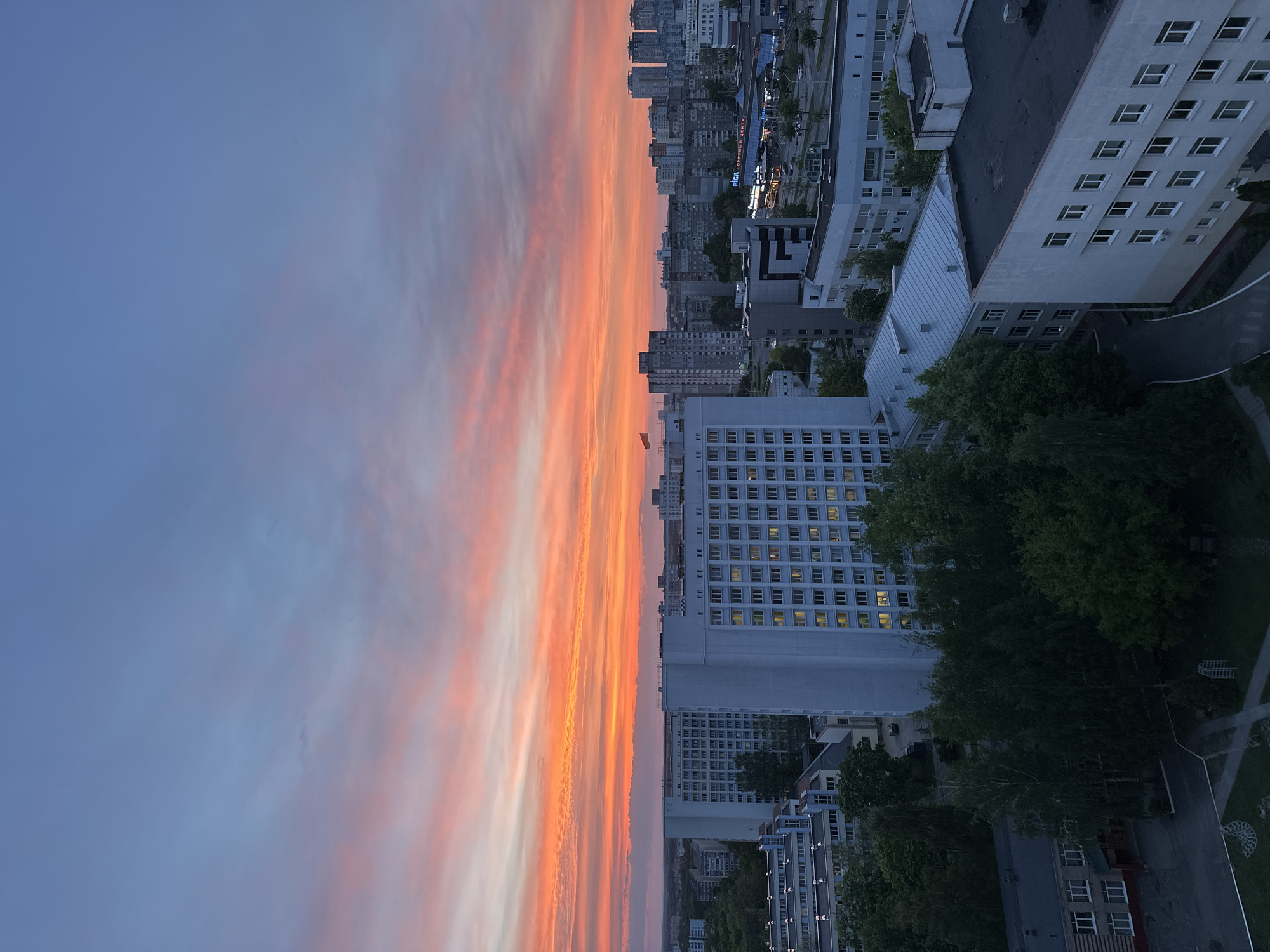 The image depicts an urban scene at twilight, likely in Minsk, Belarus, based on architectural styles. The perspective is an elevated view looking across the city, with a dramatic sunset dominating the left half of the frame. The sky exhibits a gradient from blue to orange and pink, indicating the sunset. The right side of the image contains a dense cluster of buildings, including high-rise apartments and commercial structures. There are numerous multi-story buildings with repetitive window patterns, interspersed with green spaces, suggesting parks or tree-lined streets. A large white structure with many windows is prominent among the buildings. A darker, flat-roofed building is also visible, partially obscured by trees. The overall environment suggests a densely populated area transitioning from day to night.