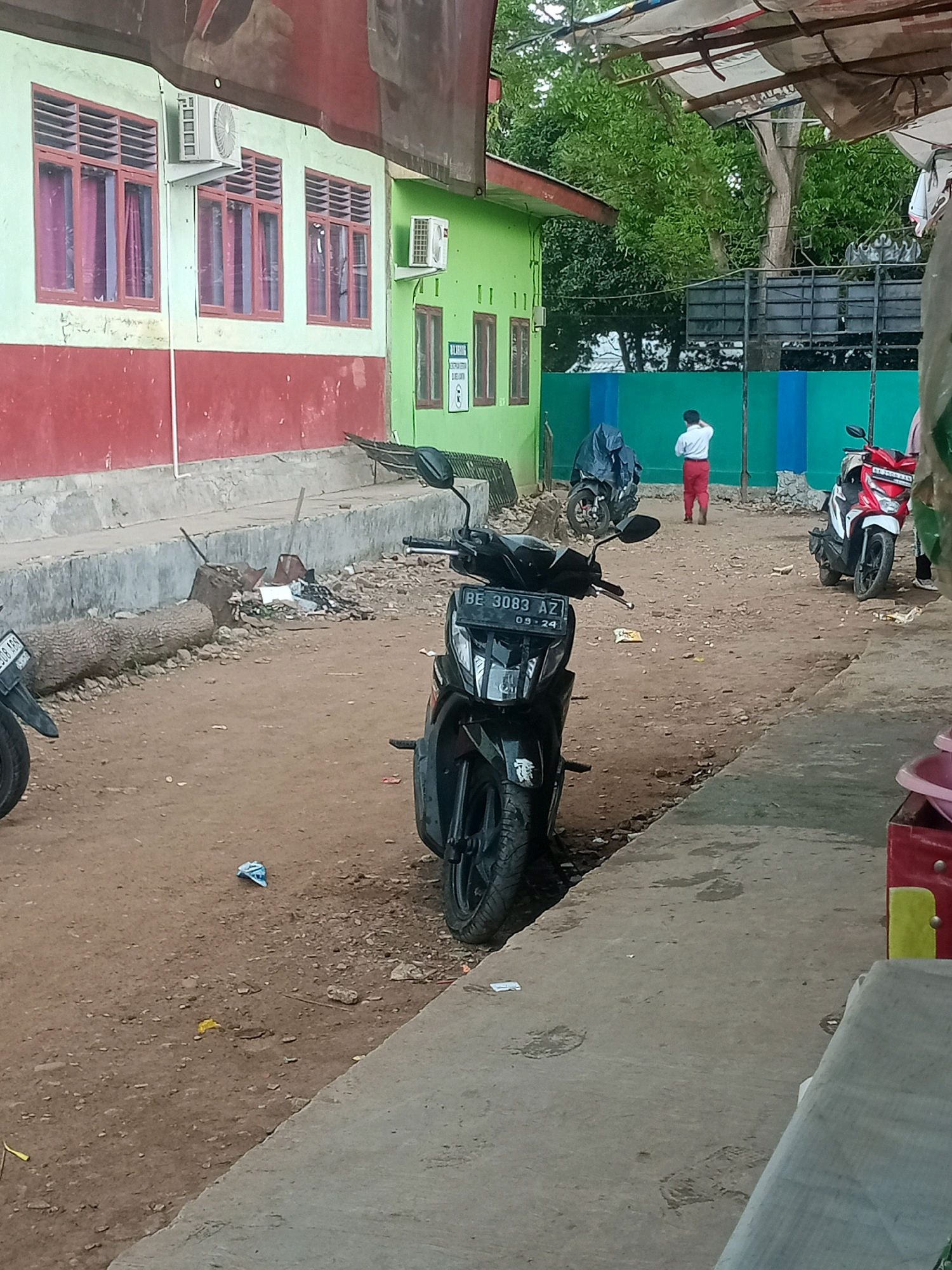 The image depicts an outdoor scene in Bandar Lampung, Indonesia, likely a schoolyard or a similar institutional setting. A two-story building with a red and light green facade dominates the background, featuring rows of windows with maroon-colored frames and louvers. Air conditioning units are mounted on the exterior walls. In the foreground, a black motorcycle with a license plate reading "BE 3083 AZ 09-24" is parked on a dirt and gravel path. To the right of the motorcycle, a red and white scooter is also visible. A person, possibly a student, wearing a white shirt and red pants, stands in the middle ground, facing away from the camera and touching their head. The environment appears somewhat disheveled, with a fallen tree trunk, scattered debris, and a blue plastic bag on the ground. Overhead, a makeshift canopy of tarps and poles provides shade. The overall impression is one of a functional, yet somewhat unkempt, public space.