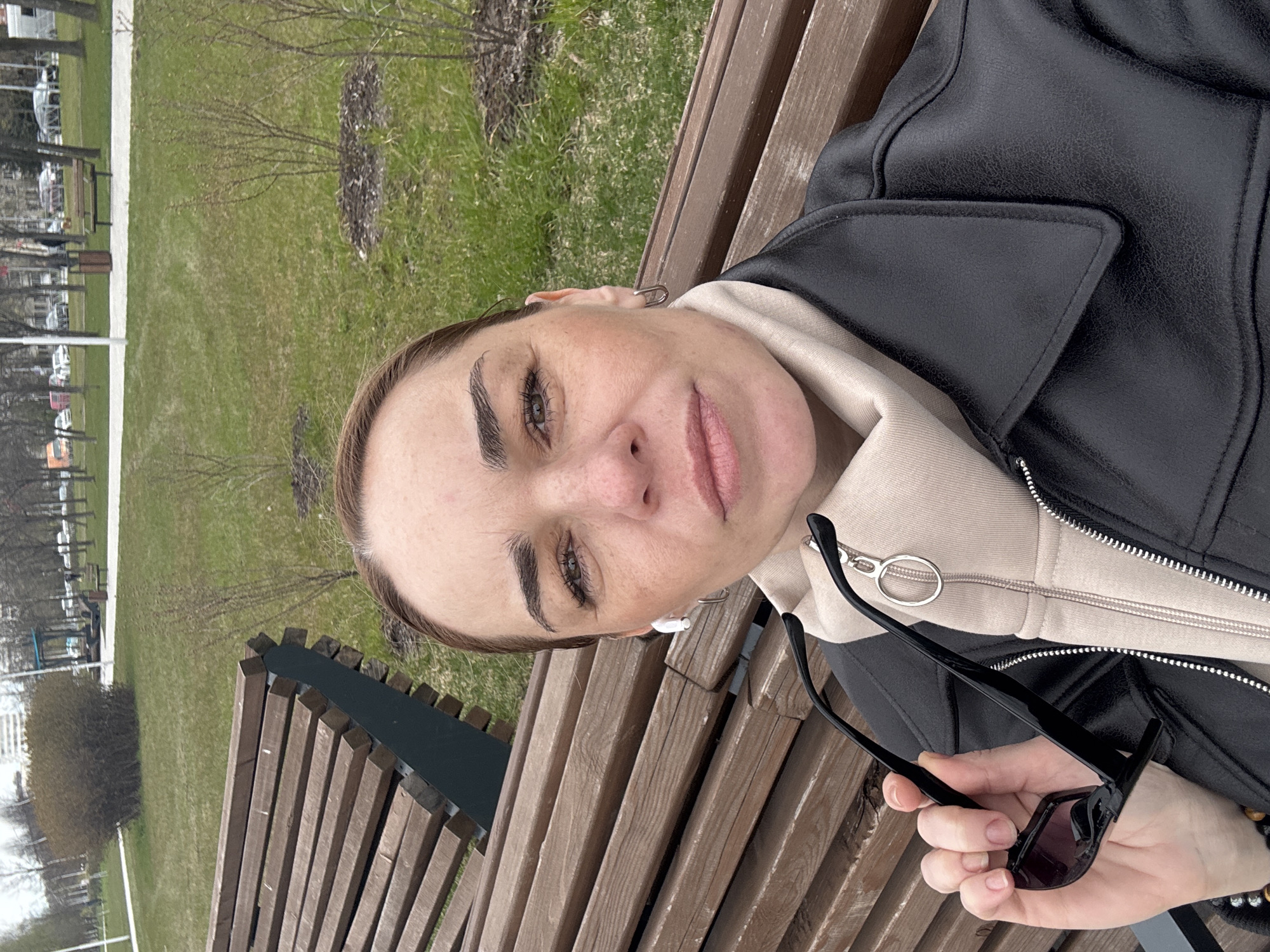 A medium close-up shot shows a person reclining on a wooden park bench in Vsevolozhsk, Russia. The person, likely a woman, faces the camera, holding sunglasses in her right hand. She wears a black leather jacket over a beige hoodie. The park setting includes grass and trees in the background, suggesting a public park or recreational area. The bench is made of light-colored wooden slats, typical of park furniture.