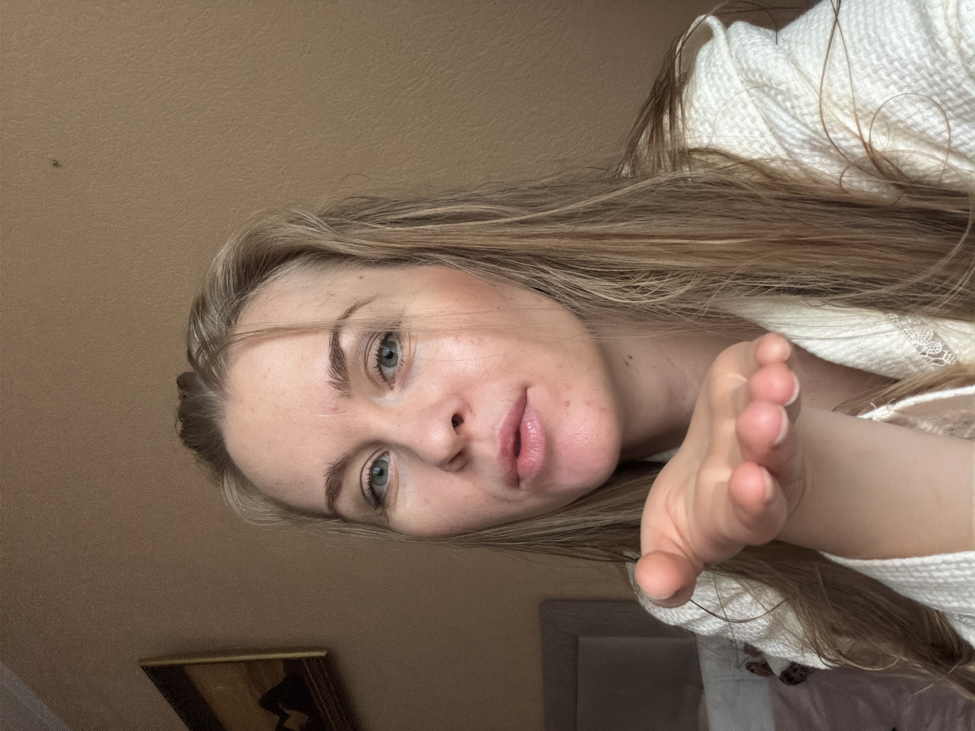 A young woman with blonde hair and blue eyes is shown in a close-up, head-and-shoulders shot. She is wearing a white textured robe and is blowing a kiss towards the camera, with her right hand extended forward. The background consists of a plain, light brown wall with a framed picture hanging on it to the left. To the lower left, a portion of a bed with pink bedding is visible. The overall lighting suggests an indoor setting, possibly a bedroom. There are no discernible features that place this image specifically in Khabarovsk, Russia.