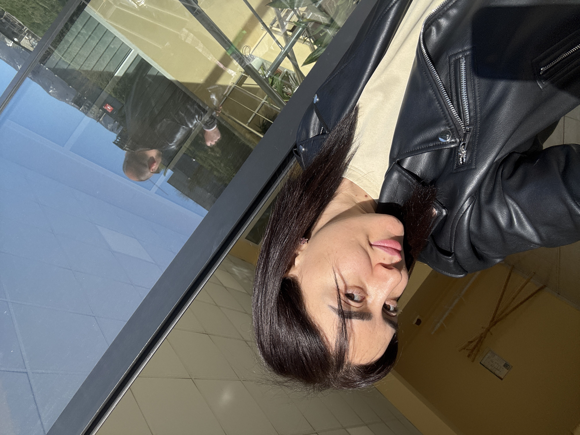 A woman with dark hair, wearing a black leather jacket and a beige shirt, poses for a selfie. Her reflection is visible in the window next to her, where a bald-headed man in a similar leather jacket can be seen. The location appears to be a building with a tiled wall, potentially in Tbilisi, Georgia, as indicated by the reflection of the outside environment visible in the window. The background shows a green area with buildings and what appears to be a mountain range. The lighting suggests it is daytime.