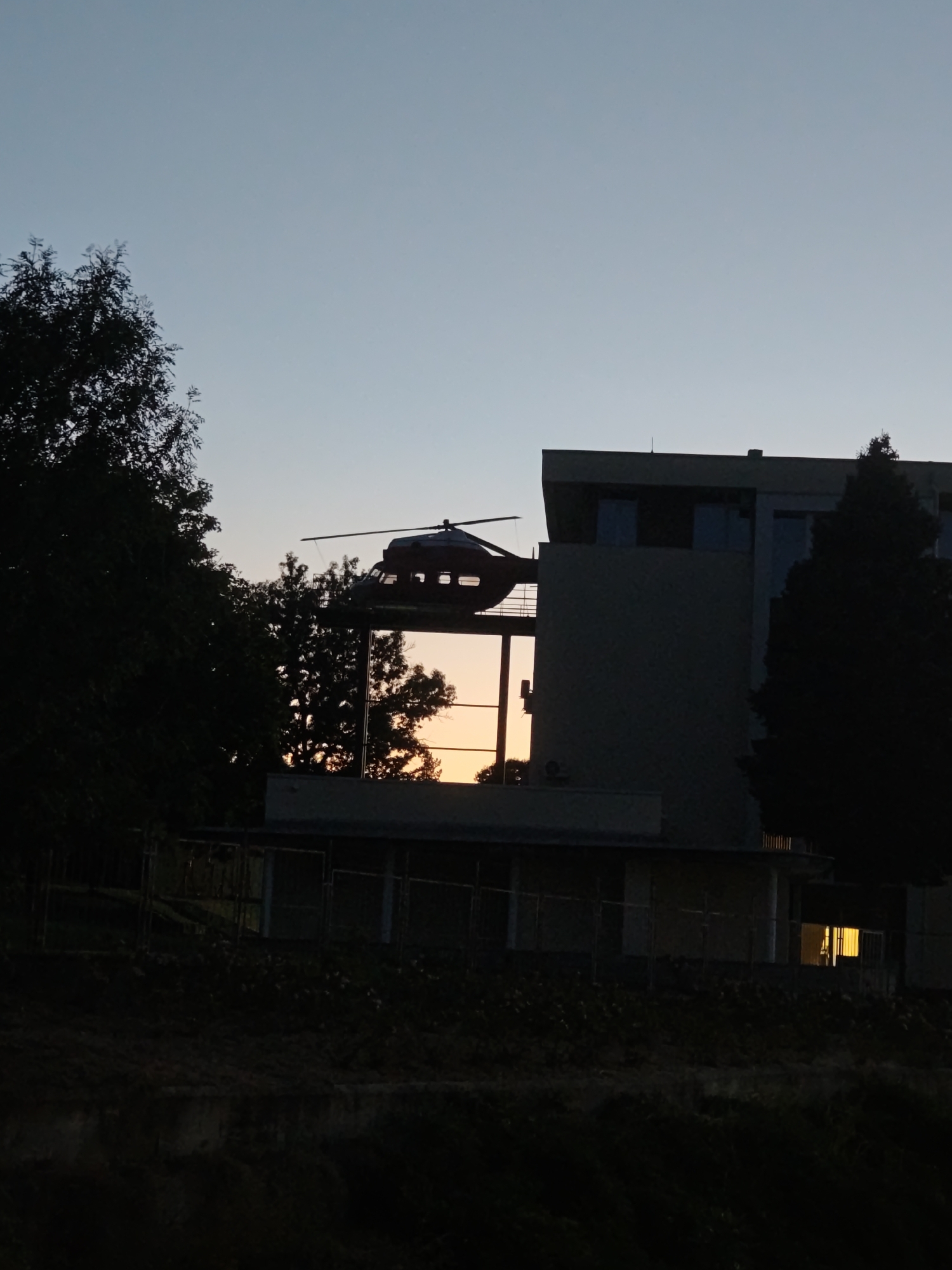 A dark silhouette of a helicopter is visible on a rooftop helipad. The helicopter is positioned in front of a building with large windows that reflect the orange and yellow hues of a sunset. Trees frame the scene on the left and right, their dark forms contrasting with the twilight sky. The image appears to be taken at dusk or dawn.