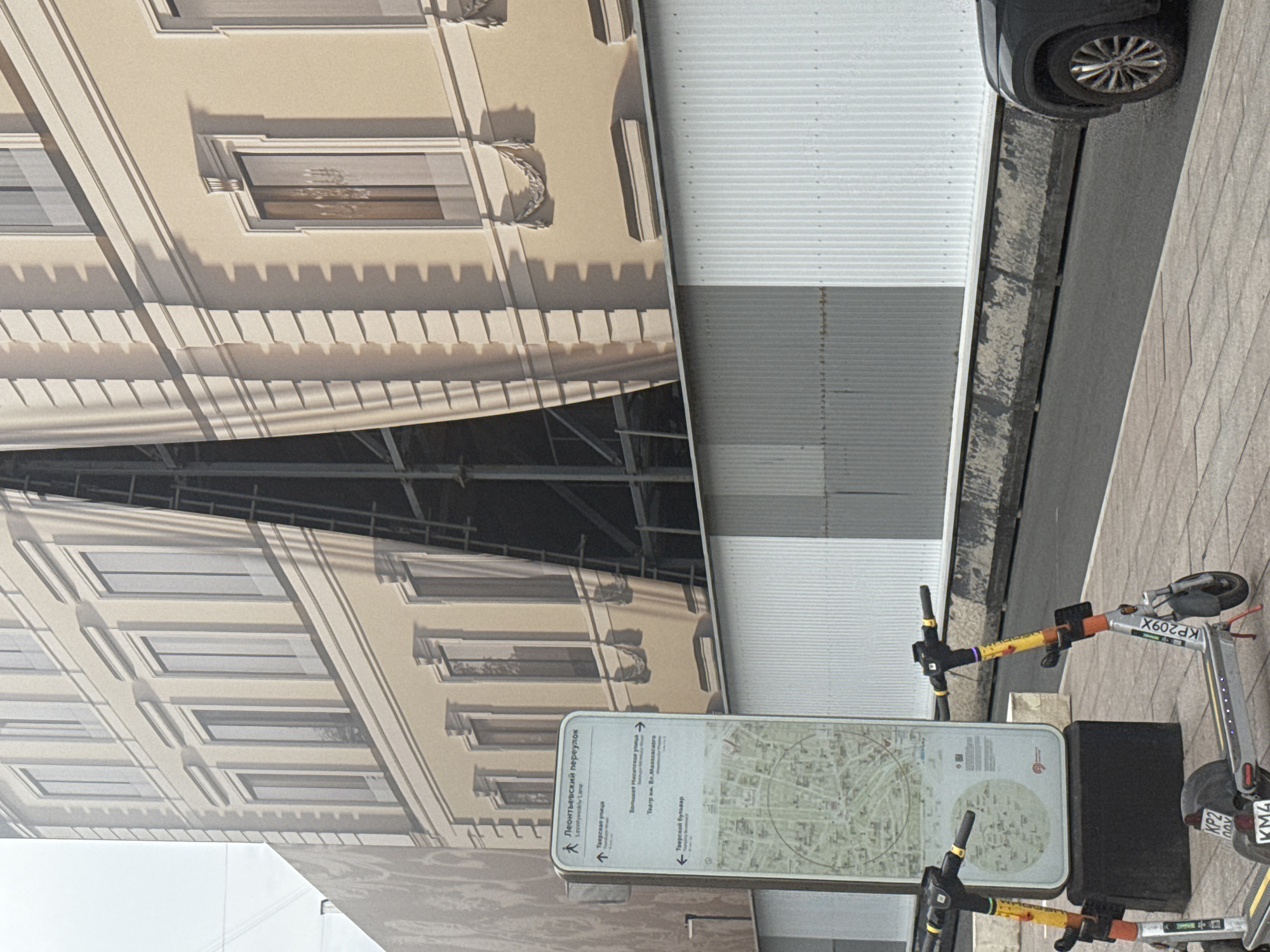 The image depicts a street scene in Moscow, Russia. A large, painted tarp covers a building under construction, simulating the facade of a historical building with windows. In the foreground, a signpost displays information in Russian, including directions to Leontyevsky Lane (Леонтьевский переулок), Tverskaya Street (Тверская улица), Bolshaya Nikitskaya Street (Большая Никитская улица), and Tverskoy Boulevard (Тверской бульвар). A map of the area is also visible on the sign. Two electric scooters are parked near the signpost, and a dark-colored car is partially visible on the right side of the image. The environment appears to be an urban street with a sidewalk and a road.