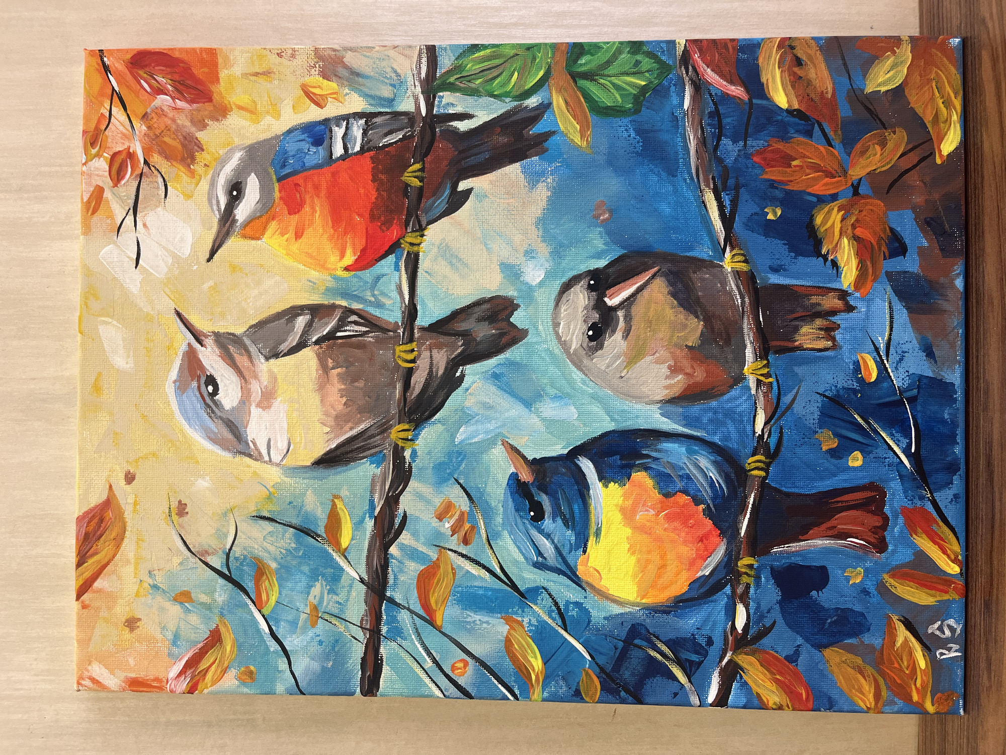 An acrylic painting depicts four colorful birds perched on branches amidst autumn leaves. The artwork has a vibrant, expressive style with visible brushstrokes, showing a mix of warm and cool tones.
The primary subjects are four small birds with varying plumage.
* The top bird, oriented horizontally, has a gray head, a blue back, and a bright orange chest and belly.
* Below it, another bird is positioned horizontally, with a lighter gray head, brown back, and a pale yellow to light brown chest.
* On the right side, a bird is oriented upside down, appearing to cling to a branch. It has a dark cap, gray face, and a mottled brown and cream body.
* At the bottom, another bird is depicted upside down, with a dark blue head, blue back, and a vivid orange and yellow chest/belly.
The birds are resting on dark brown, thin branches. Scattered around them are numerous leaves in shades of orange, red, and yellow, characteristic of autumn foliage. The background is divided into two main color schemes: the left side features lighter blues and yellows, while the right side is dominated by deeper blues. This contrast in the background colors adds dynamic interest to the composition.
There is no specific activity or event taking place other than the birds perching. The scene conveys a sense of autumn with its warm-colored leaves. The bright lighting suggests it is daytime. The painting's style is painterly, with thick, textured applications of paint evident, especially in the background and on the leaves.
In the bottom right corner, a signature "RS" is visible. While the painting depicts a generic nature scene, it is associated with Burgas, Bulgaria.