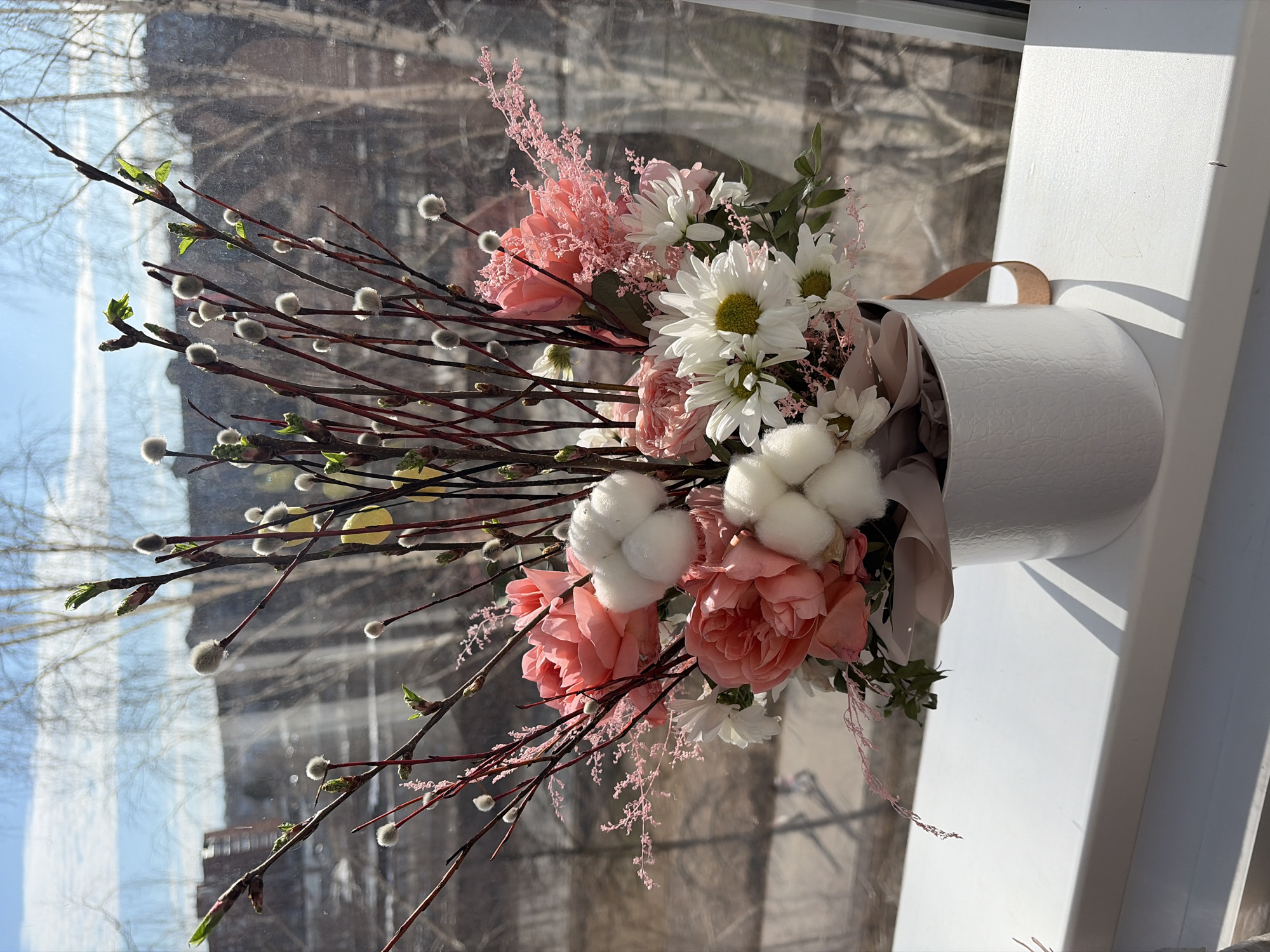 Here is a forensic description of the image:
**Scene:** A bouquet of flowers and branches arranged in a white cylindrical container. The arrangement is positioned on a white windowsill, against a window with an obscured view of an outdoor scene. The scene is bathed in bright sunlight, casting shadows.
**Subjects:**
* A bouquet composed of pink roses, white chrysanthemums, cotton flowers and pussy willow branches.
**Objects:**
* White cylindrical container with a textured pattern.
* Tan ribbon looped around the container.
* White windowsill.
* Window with partially visible outdoor scene.
**Environment:**
* The outdoor scene visible through the window is blurry but appears to show trees without leaves and buildings.
**Assumptions based on visual clues:**
* The location is Chaikovsky, Russia based on the image description.
* The time of year is likely late winter or early spring, given the presence of budding branches and limited foliage.
