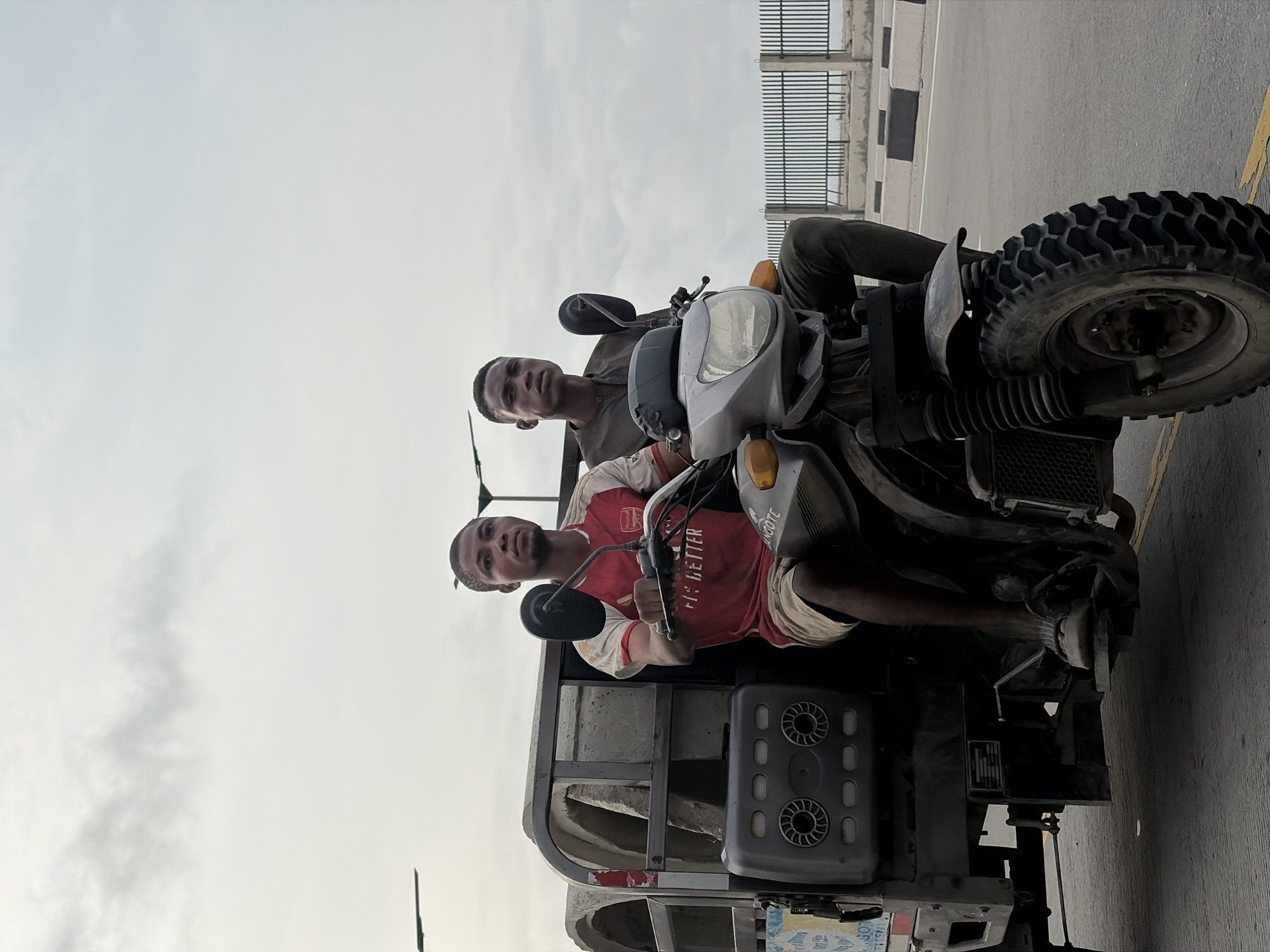 In Alaguntan, Nigeria, under a cloudy sky, two young men are seen riding a motorbike that is attached to a cargo carrier. The man in front, the driver, is wearing a red and white Arsenal football jersey and khaki shorts, and he is holding the handlebars. Beside him, the passenger is wearing a dark grey shirt and dark pants. The motorbike has a large, rugged tire and the cargo carrier behind it contains several large, cylindrical concrete objects. The scene appears to be on a paved road, with a metal fence and more concrete barriers visible in the background. Streetlights are also present in the distance.