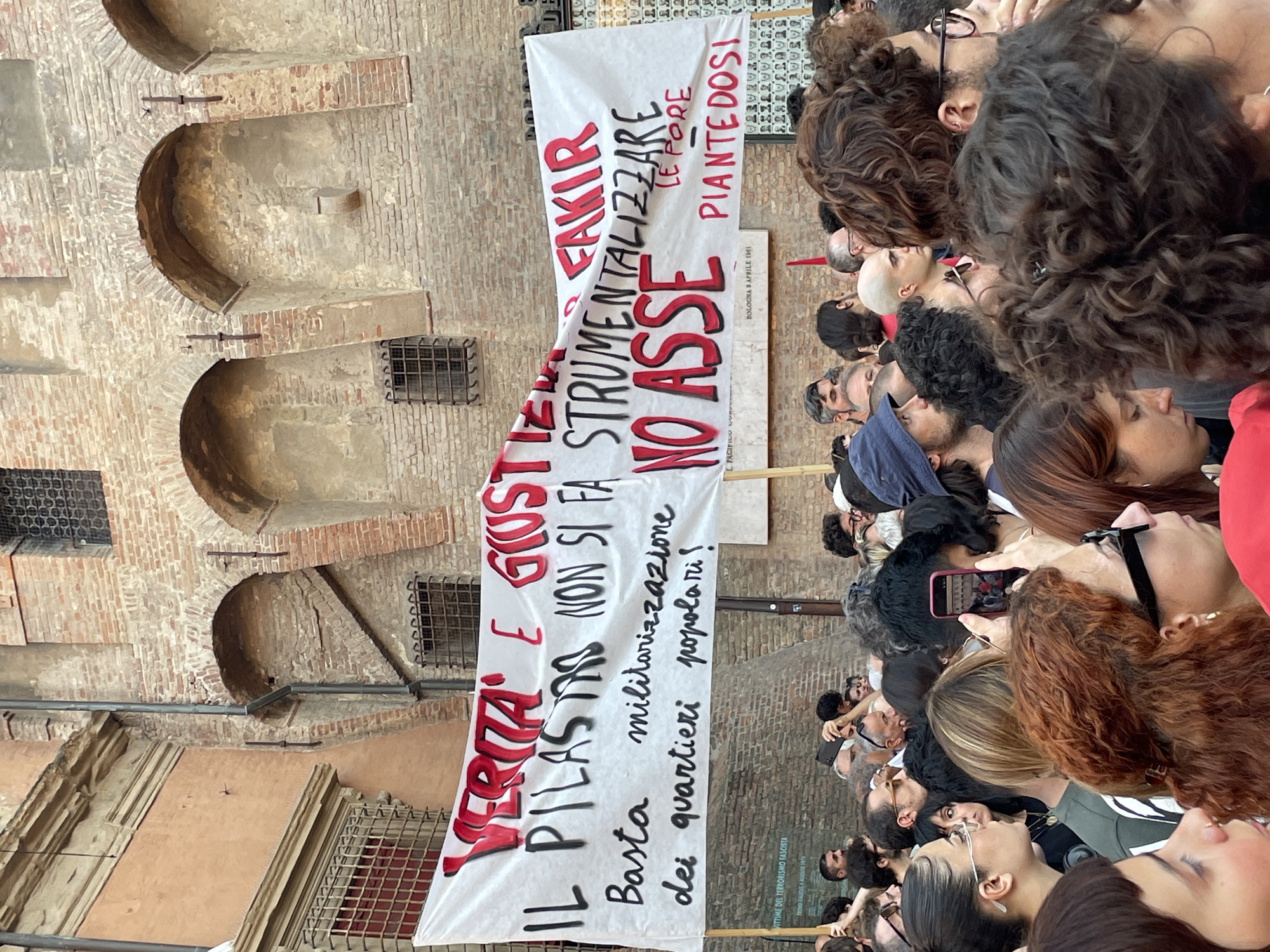This media file shows a crowd of people gathered outdoors, with a banner displayed prominently. The banner, written in Italian, reads: "VERITA E GIUSTIZIA IL PILASTRO DELLA STRUMENTALIZZARE NON SI FA BASTA militarizzazione DEI QUARTIERI POPOLARI! LE PORE PIANTEDOSI". It also includes the text "BOLOGNA 9 APRILE 1961" and "VITTIME DEL TERRORISMO FASCISTA". The scene appears to be in a historical urban setting, with old brick buildings featuring arched windows and openings. The people in the crowd are looking upwards, seemingly at the banner or something beyond the frame. Their expressions are varied, with some appearing serious or pensive. The lighting suggests it is daytime, likely bright and sunny. The overall atmosphere seems to be one of a protest or demonstration. The crowd is dense, with many individuals' faces visible, including a bald man in the center. The banner is held up by two poles.