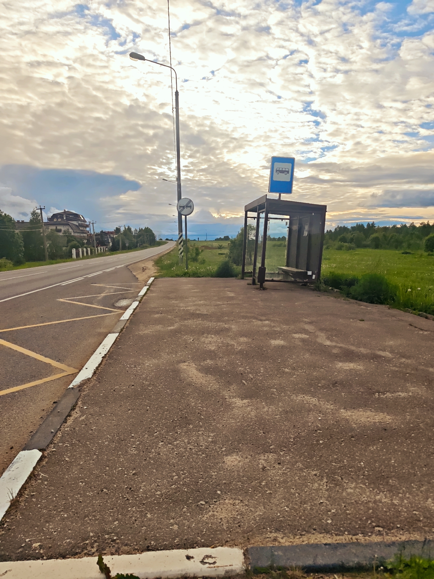 The image shows a roadside scene in Курово, Russia. A bus stop shelter with a blue bus stop sign is situated next to a paved area adjacent to a two-lane asphalt road. The road has yellow no-parking markings. A lamppost and a road sign are visible further down the road. In the background, there is a green field and forested area. There is a building set further back from the road. The sky is cloudy. There are no people visible in the image.