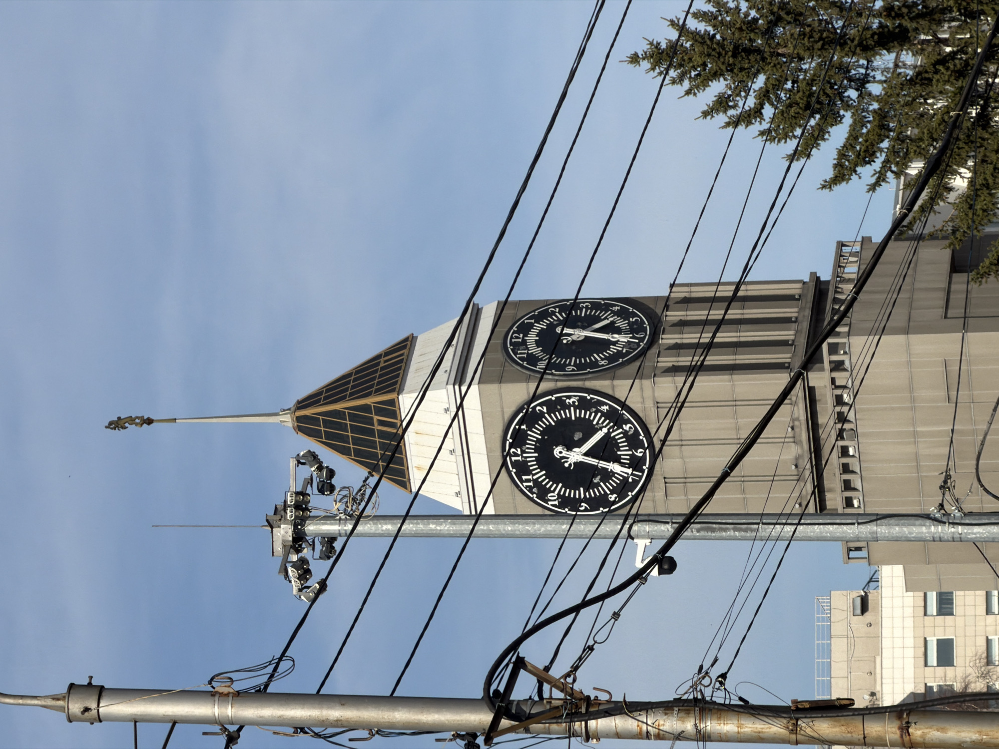 The image depicts the Krasnoyarsk Clock Tower against a blue sky. The clock tower is a tall, light-colored building with two large, round clocks visible on its facade. A pyramidal structure tops the tower. Power lines crisscross the foreground. A utility pole runs vertically in the lower center of the frame. Part of a tree is visible in the top right corner. A building with several windows is visible on the bottom right.