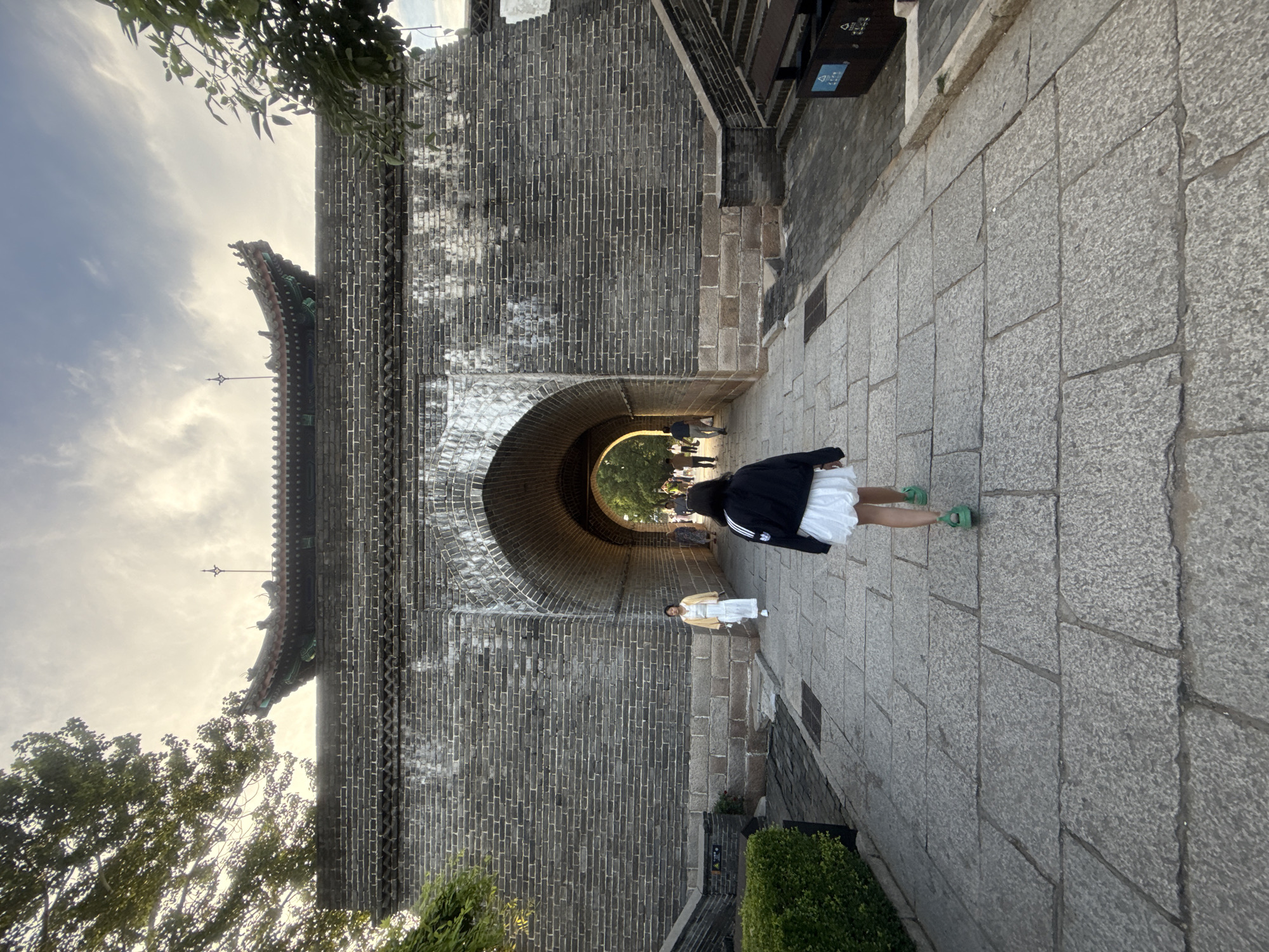 The image shows a person walking towards a large, ancient brick gate in Qinhuangdao, China. The gate is made of dark gray bricks and has a large arched opening. Above the arch, there is a decorative roof with green tiles and ornate carvings. The path leading to the gate is paved with large, flat stones. The sky above is cloudy with some sunlight breaking through.
To the left of the path, there are green bushes and a stone staircase leading up. To the right, there is another stone staircase and a trash bin. Inside the archway, other people can be seen walking in the distance, and greenery is visible beyond the gate.
The person in the foreground is wearing a black jacket with white stripes on the sleeves and a white skirt. They are also wearing green shoes. The person is walking away from the camera, towards the gate. The overall atmosphere of the image suggests a historical site or attraction within the city of Qinhuangdao.