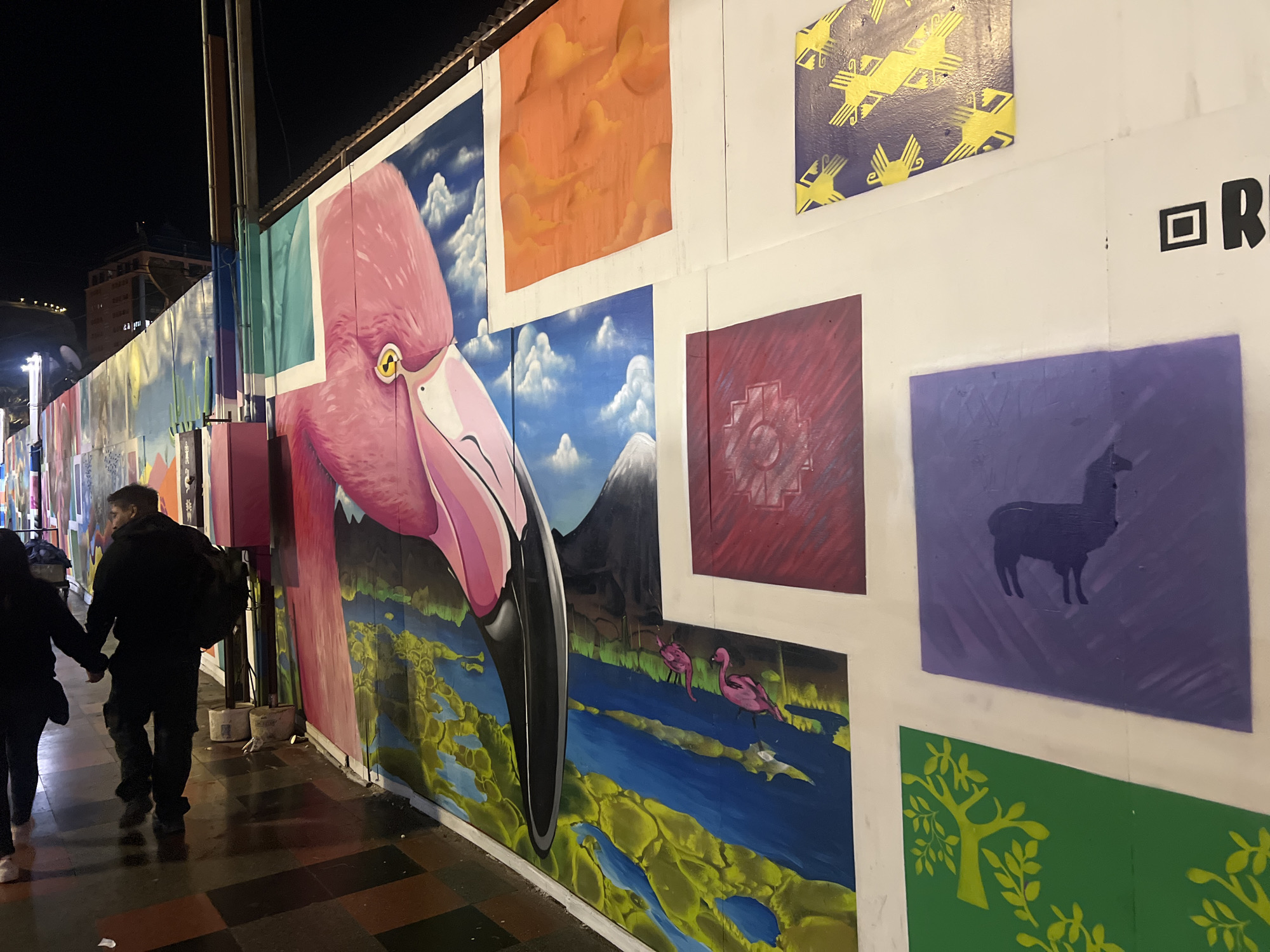 This image captures a vibrant street art mural located in Arica, Chile. The mural features a large, close-up depiction of a pink flamingo's head and beak, set against a backdrop of a blue sky with white clouds and distant mountains. To the right of the flamingo, a series of smaller, colorful panels display various artistic motifs: a square of abstract orange clouds, a purple square with a silhouette of a llama, and a green square with a stylized yellow tree. Further to the right, a black graphic appears to be part of a larger piece of text or signage. In the foreground, two individuals, a man and a woman, are walking hand-in-hand along a wet, tiled sidewalk. They are both dressed in dark clothing, with the man carrying a backpack. The overall scene is illuminated by artificial light, suggesting it is nighttime or a dimly lit area. The mural adorns a temporary construction barrier or wall, indicated by visible seams and panels.