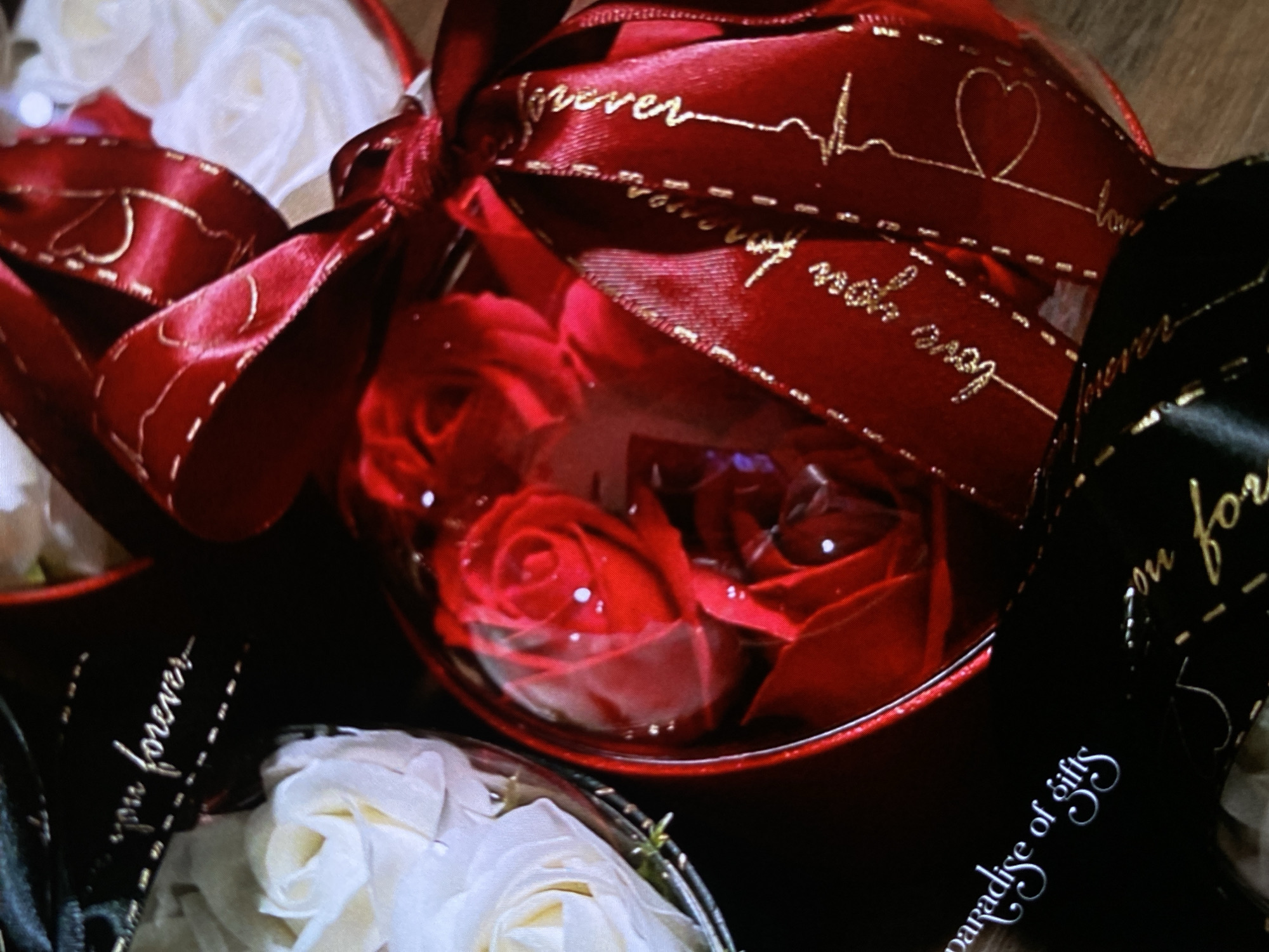 This close-up image displays an arrangement of decorative gift items, likely artificial flowers, set in clear spherical containers and adorned with ribbons. The scene focuses on several individual gift units, suggesting a display from a gift shop or a collection of presented gifts.
The central and most prominent item is a clear spherical container filled with several vibrant red artificial roses, possibly made of soap or fabric. This sphere is tied with a glossy red ribbon featuring golden accents. The design on this red ribbon includes cursive text saying "forever" and "love you," interspersed with small heart shapes and a stylized ECG (electrocardiogram) line, symbolizing a heartbeat.
To the left and bottom of the frame, parts of similar clear spherical containers are visible, holding white artificial roses. Another item to the right features a glossy black ribbon with golden cursive text. Partially visible on this black ribbon are the words "...you forever" and below that, rotated ninety degrees, the phrase "paradise of gifts."
The lighting in the image is somewhat dim, creating reflections and highlights on the clear plastic of the spherical containers. The overall impression is one of romance and gifting, with the chosen colors of red, white, and black, and the recurring romantic phrases. No people are visible. The items are neatly arranged, suggesting they are either on display or carefully packaged. The location context of Bankya, Bulgaria, is provided, but no specific visual cues in the image confirm this location beyond it being a setting for a gift display.